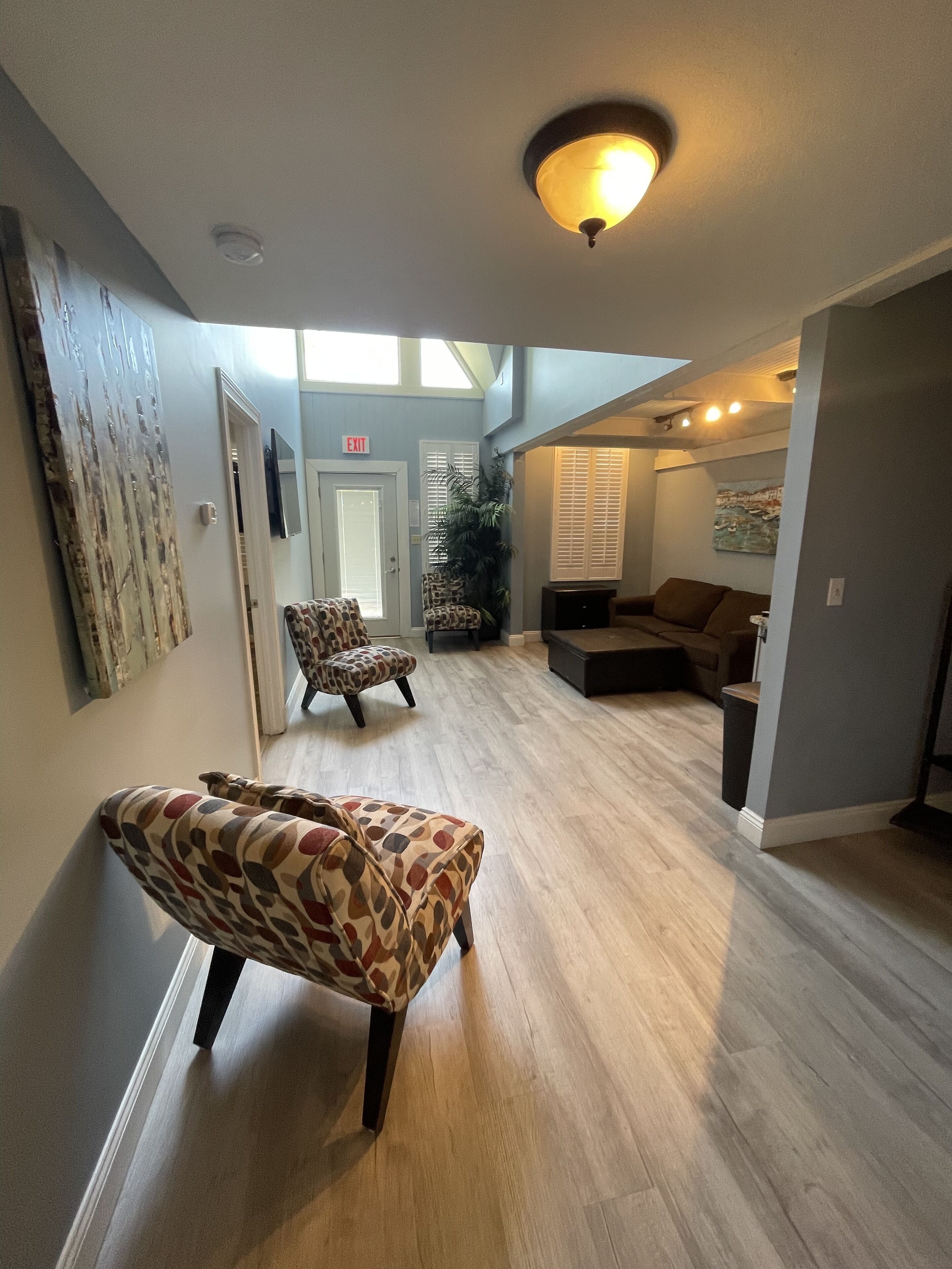 A spacious hallway with a patterned ottoman, a sofa, and a chair. There is a door with an exit sign, a plant, and artwork on the walls.