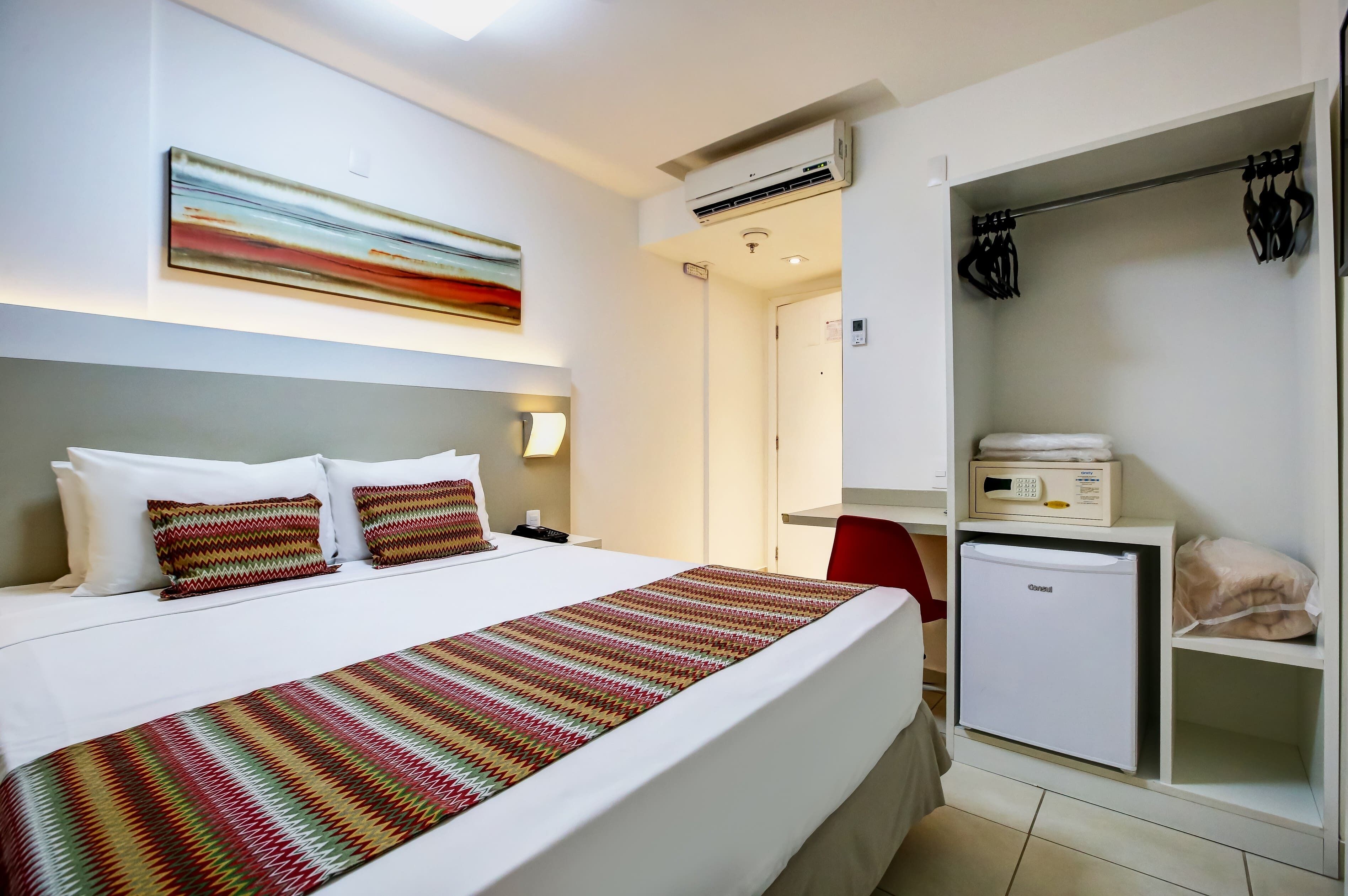A hotel room with a bed, a bedside table, a wall-mounted air conditioner, a mini-fridge, and a wardrobe.