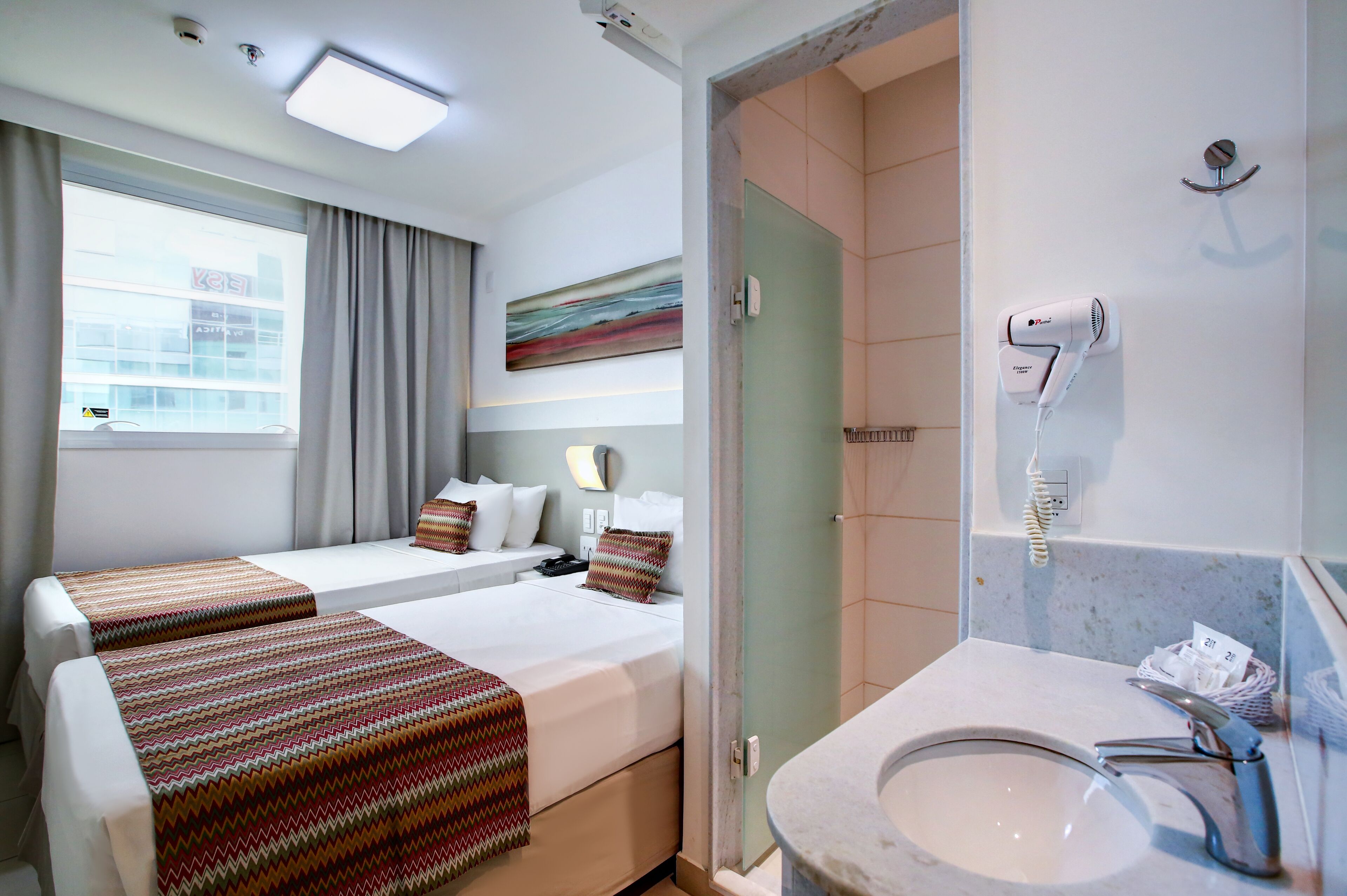 Habitación de hotel con dos camas, baño con lavamanos y secador de pelo, y ventana con cortinas.