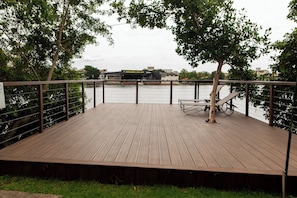 Une terrasse en bois avec des garde-corps en métal, donnant sur une rivière et offrant une vue sur des bâtiments en arrière-plan.