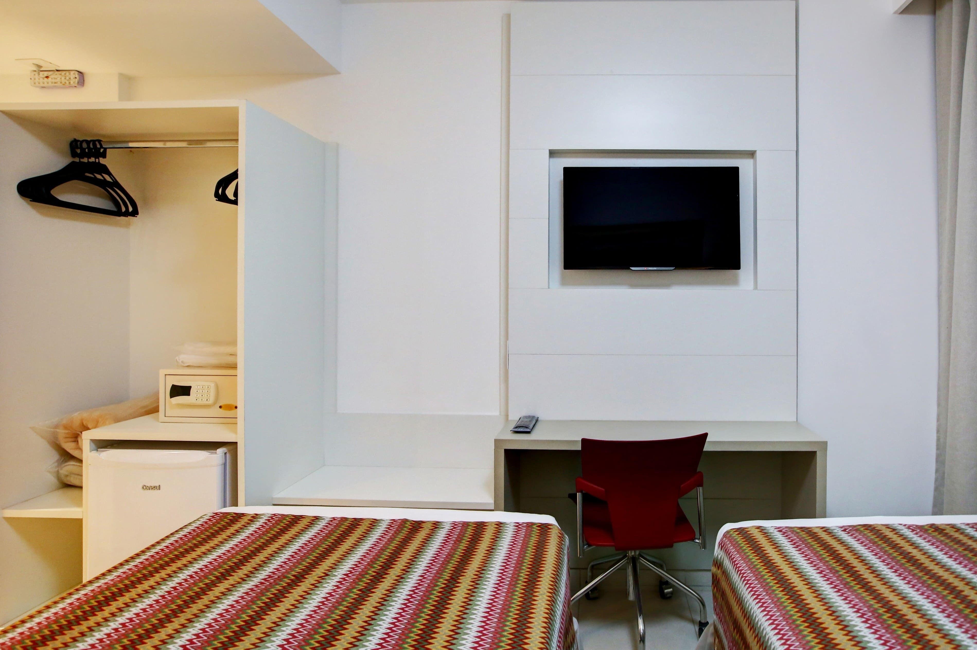 Quarto de hotel com duas camas, uma escrivaninha, uma televisão, micro-ondas e uma cadeira vermelha.
