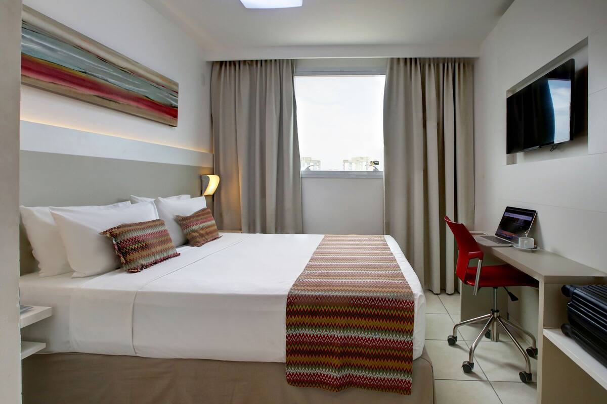 Ein Hotelzimmer mit einem Bett, einem Schreibtisch mit Laptop, einem Fernseher und einem Fenster mit Vorhängen.