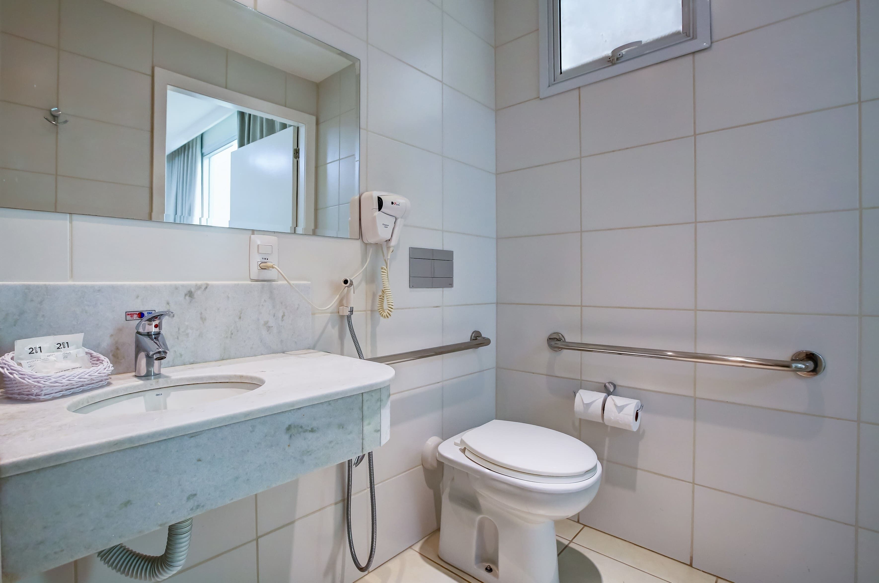 Baño con lavamanos, espejo, secador de pelo, inodoro y barras de apoyo.