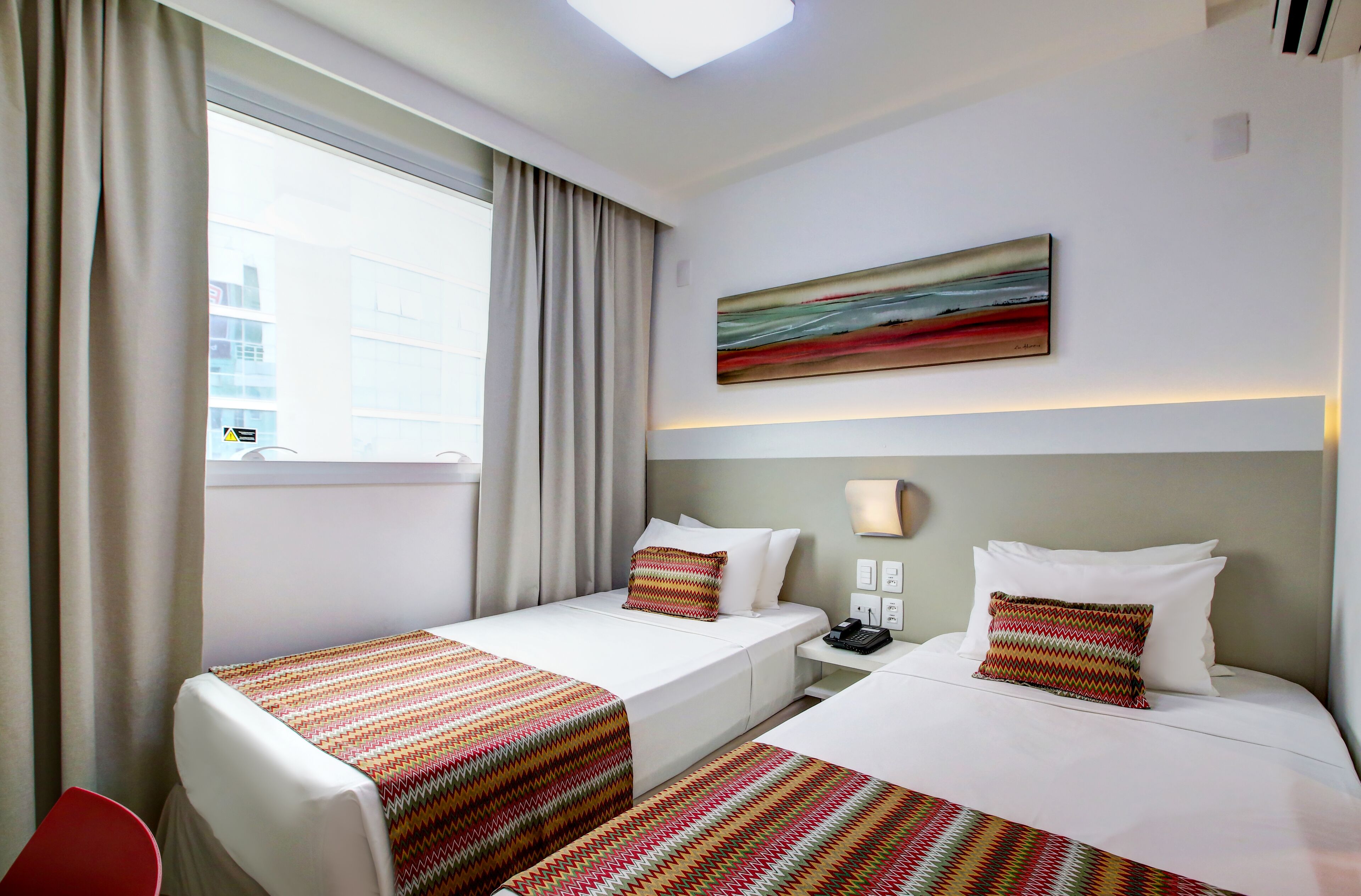 A hotel room with two beds, a window with curtains, a painting on the wall, and a desk with a phone.