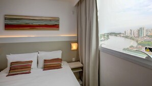 A hotel room with a bed, a painting, a nightstand, a lamp, and a window with a city view. - Transamerica Fit Vitória Reta da Penha (Vitoria)