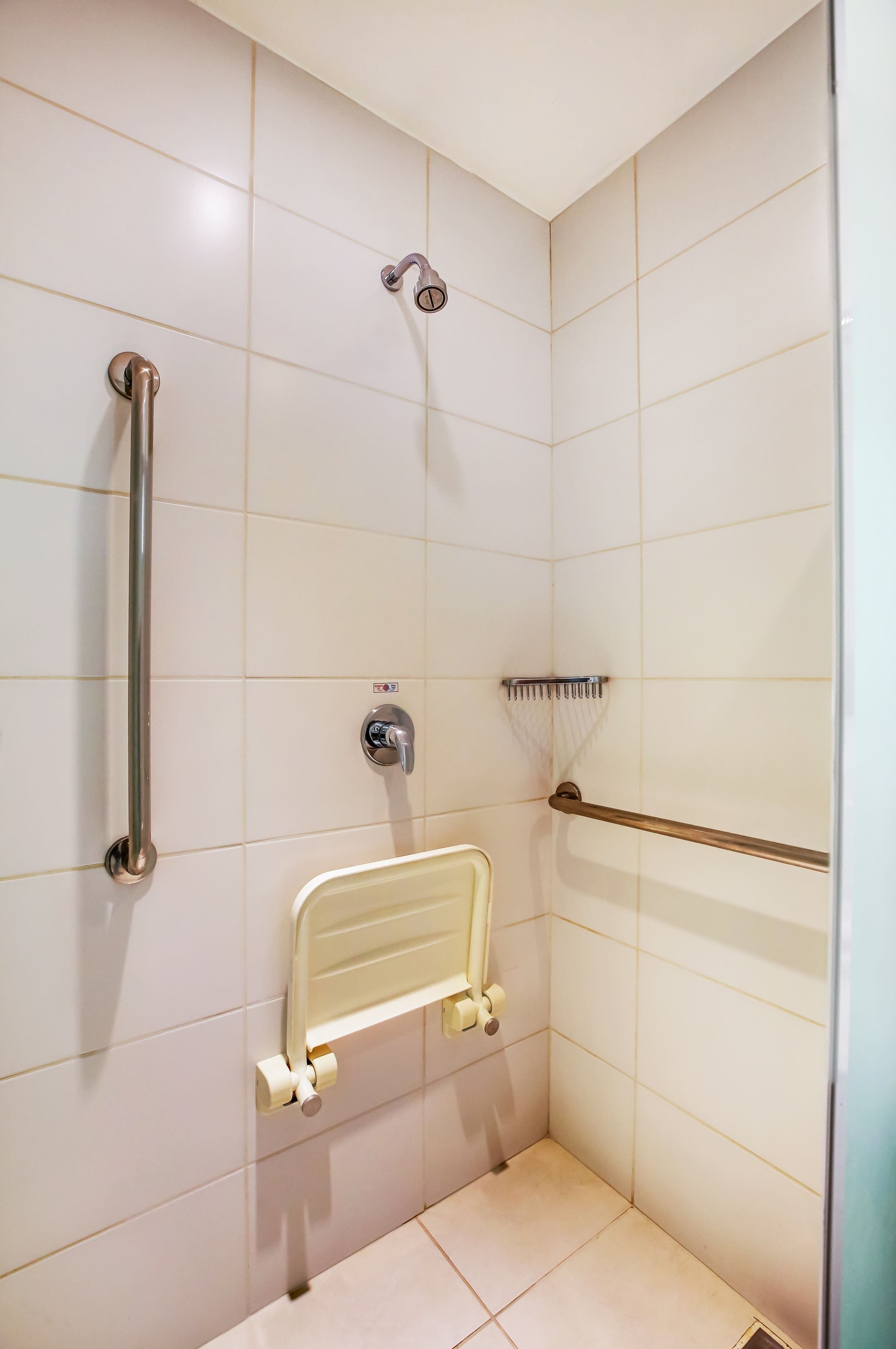 Egy fürdőszoba fehér csempékkel, kézi zuhanyfejes zuhanyzóval, kapaszkodóval, biztonsági kapaszkodóval ellátott WC-vel és kézszárítóval.