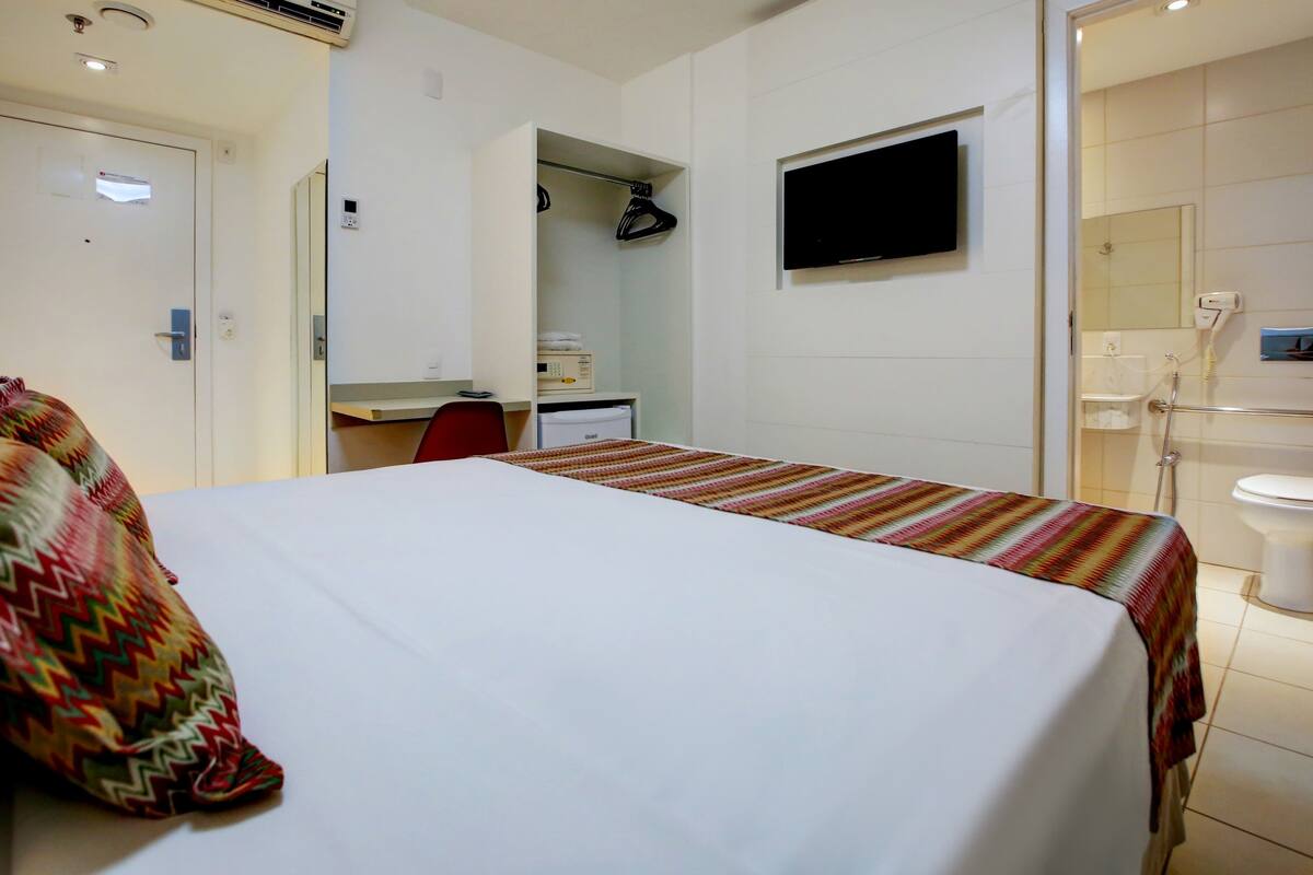 Ein Hotelzimmer mit Bett, Schreibtisch, Stuhl, einem an der Wand befestigten Fernseher und einer Dusche.