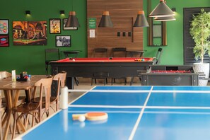 Une table de billard, une table de jeu de flipper et une table de baby-foot dans une pièce aux murs verts et aux meubles en bois.
