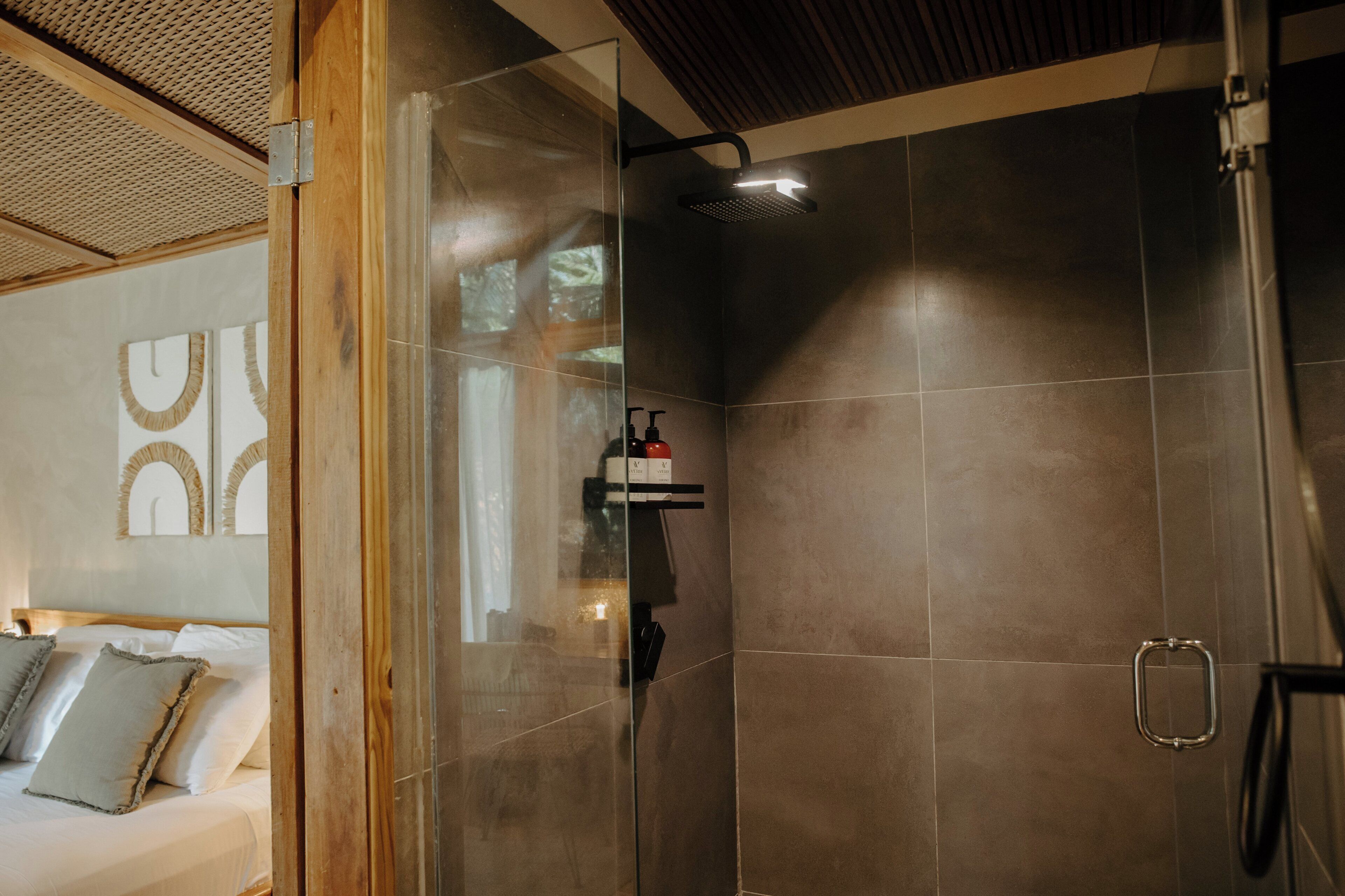 Un baño moderno con cabina de ducha de vidrio, techo de madera, y una cama con almohadillas en la habitación contigua.
