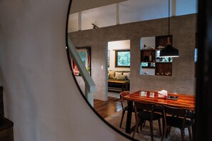 Un comedor con mesa y sillas de madera, una lámpara colgante y vista a otra habitación con sofá y ventana a través de un vano circular.