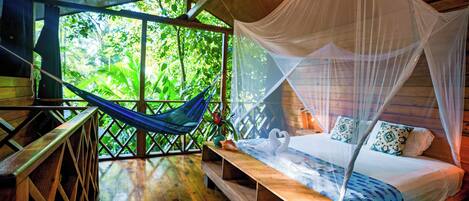 A wooden room with a bed, a hammock, and a view of lush greenery.
