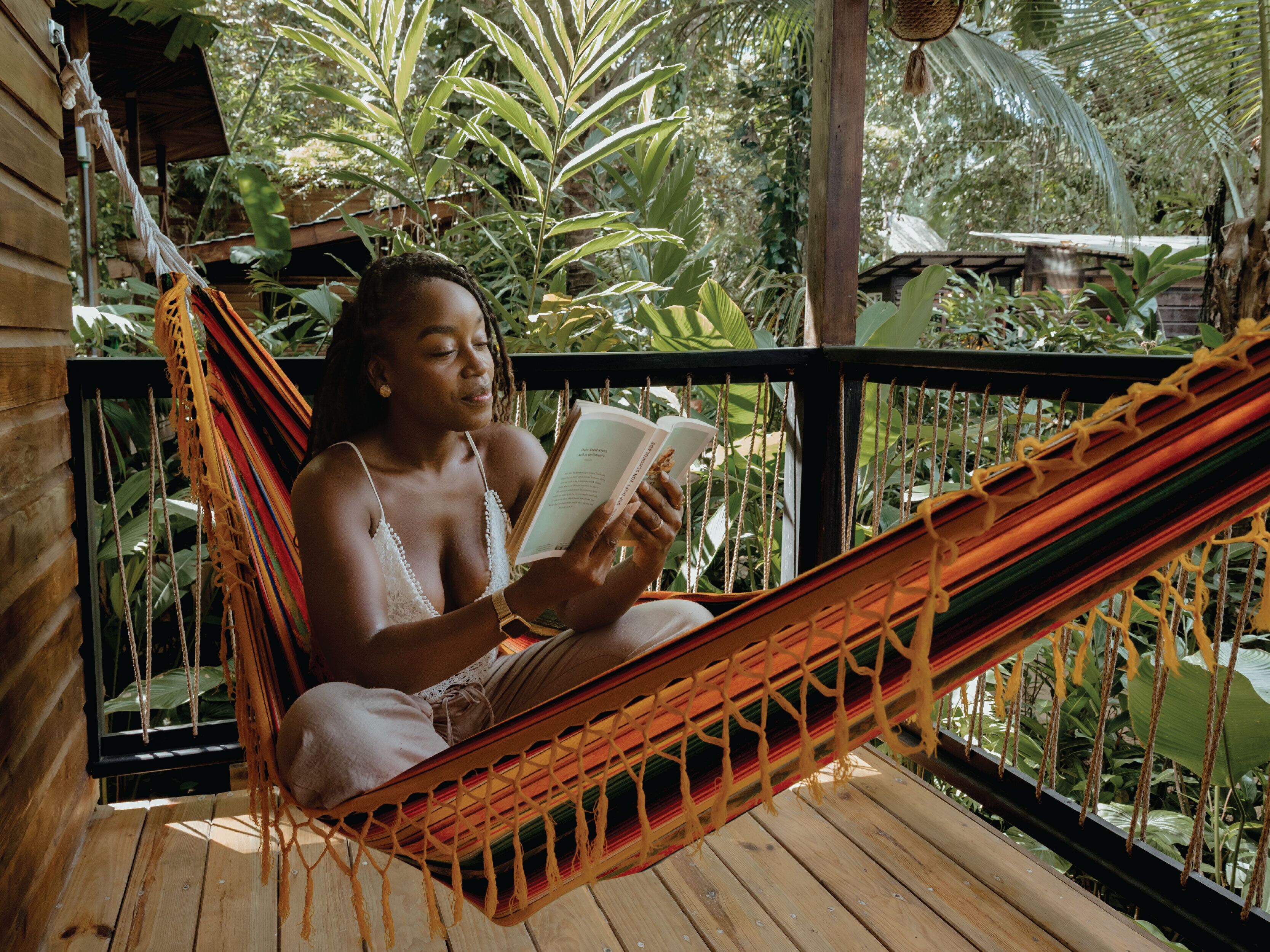 Une personne est assise dans un hamac sur une terrasse en bois, un livre à la main, avec une végétation luxuriante en arrière-plan.