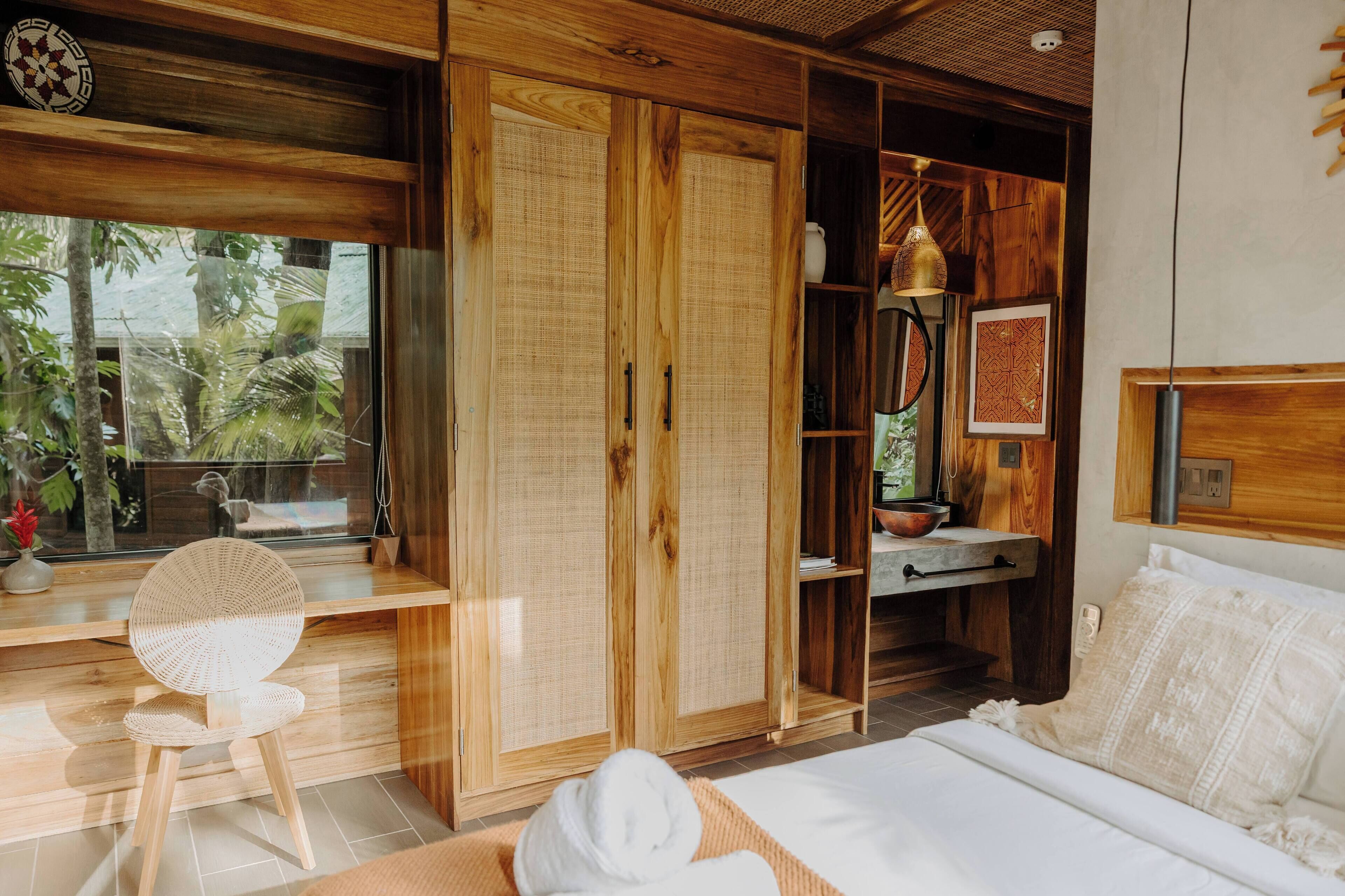 Una habitación de madera con una cama, un sillón de mimbre, un armario y una ventana con vistas a árboles.