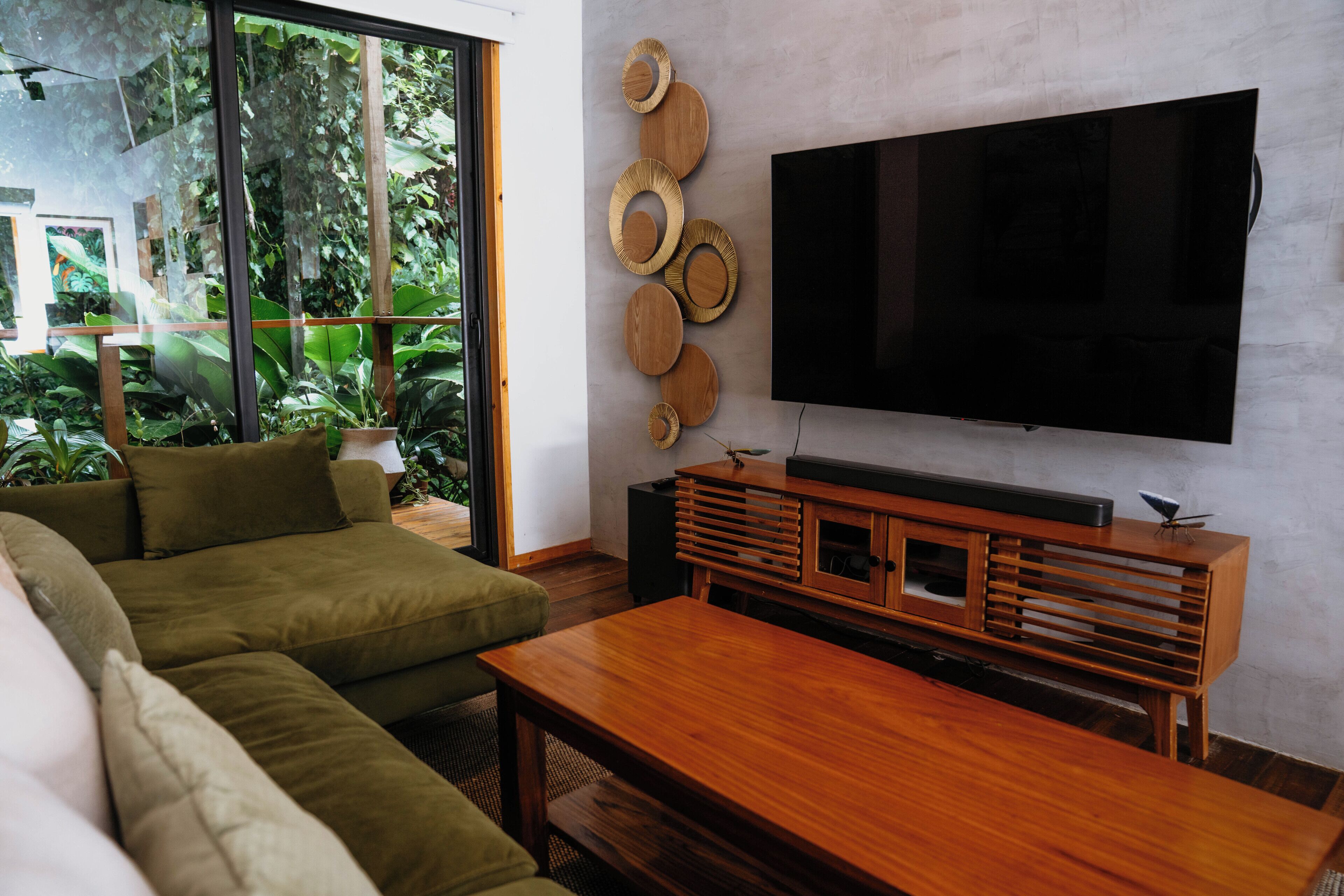 A living room with a flat-screen TV, a wooden coffee table, a green sofa, and a wall-mounted wooden shelf.
