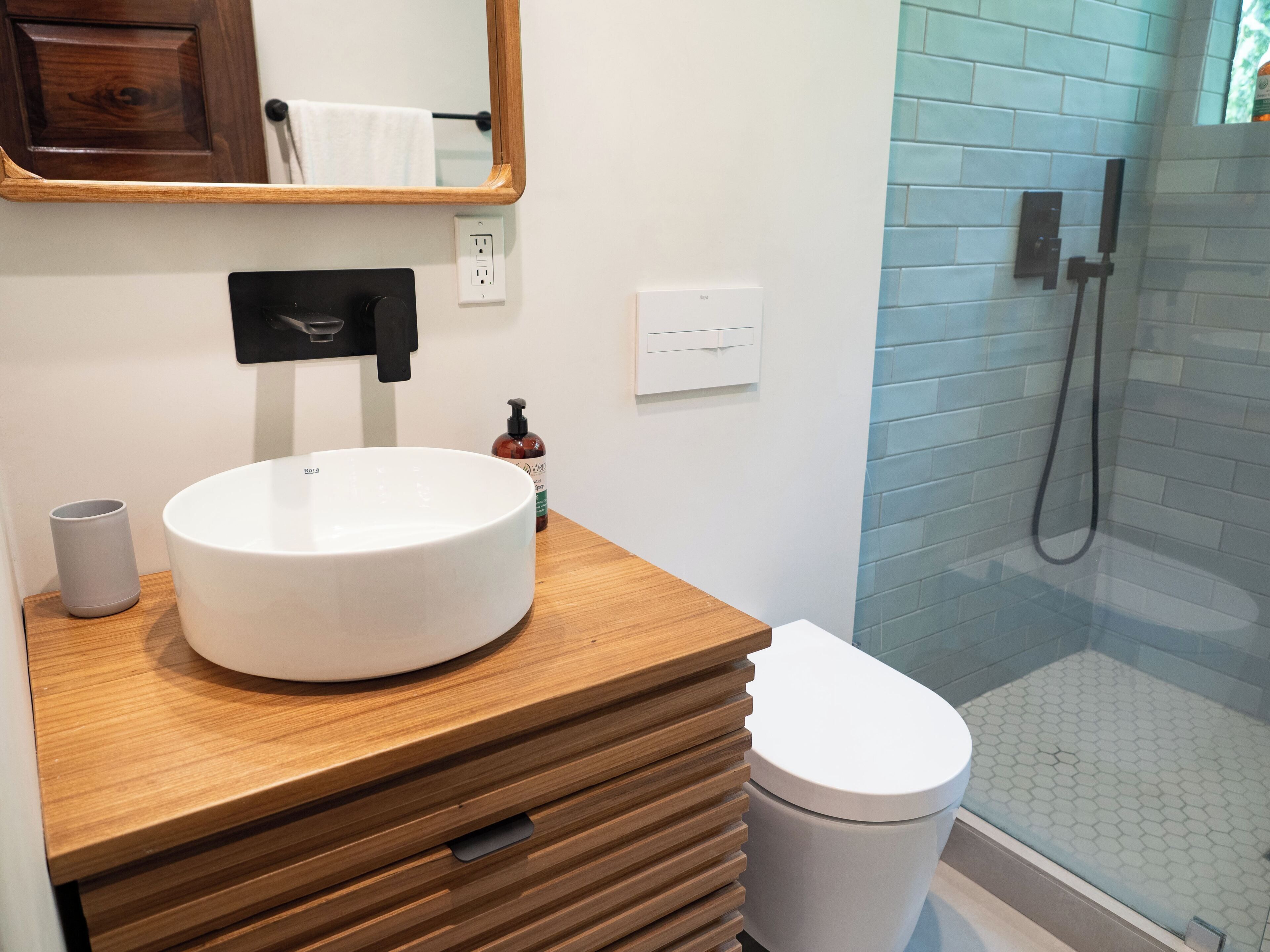 Un bagno con un mobile bagno in legno, un lavandino bianco, un water, uno specchio, un porta asciugamani e uno spazio doccia con pavimento piastrellato.