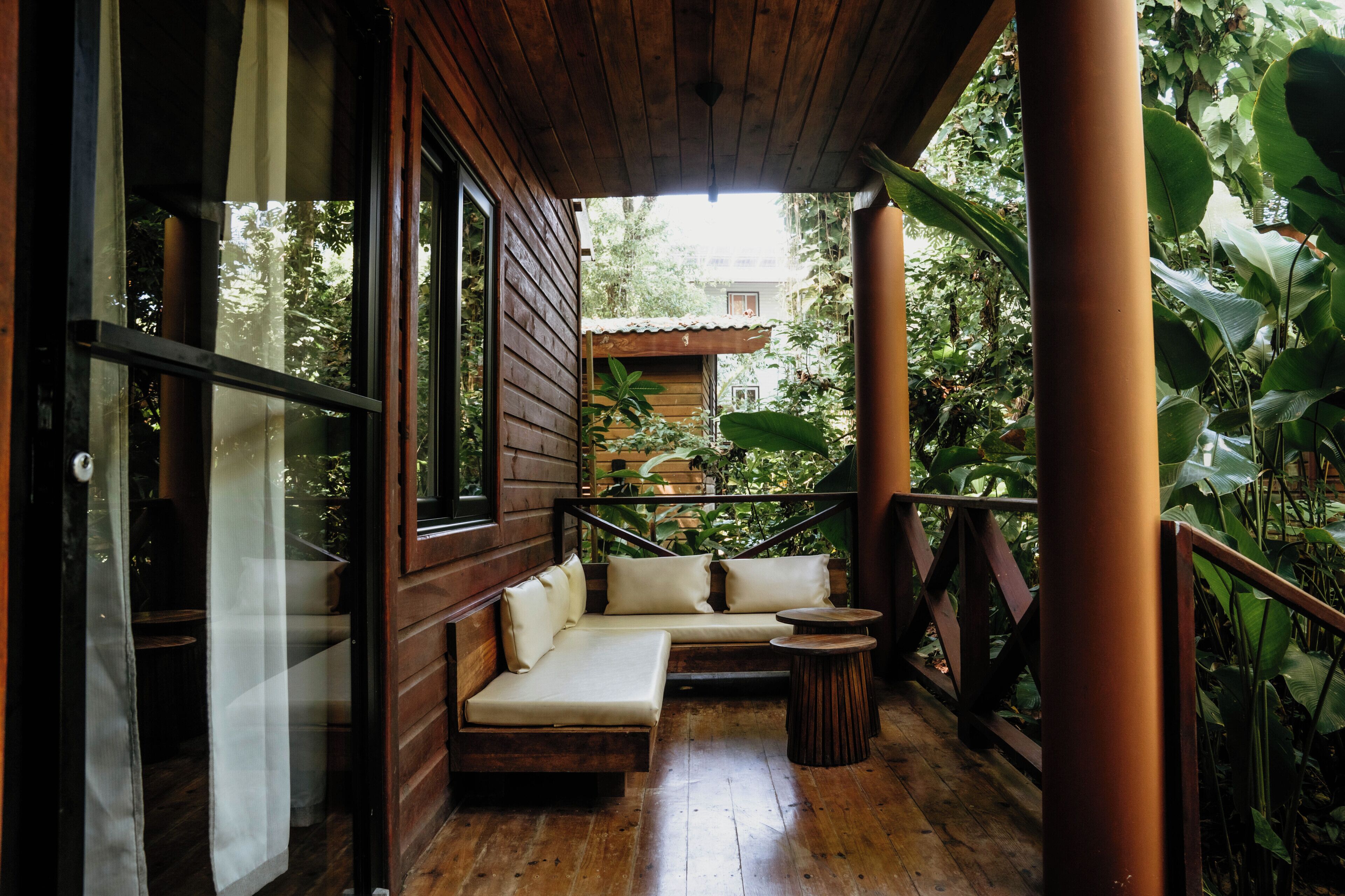 Una terraza de madera con zona de estar acolchada, una pequeña mesa redonda y vista a una casa al fondo.