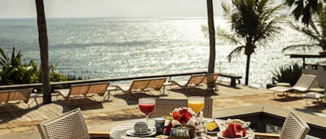Una mesa redonda con alimentos para el desayuno y bebidas, con vista a una playa con palmeras y sillas de descanso.