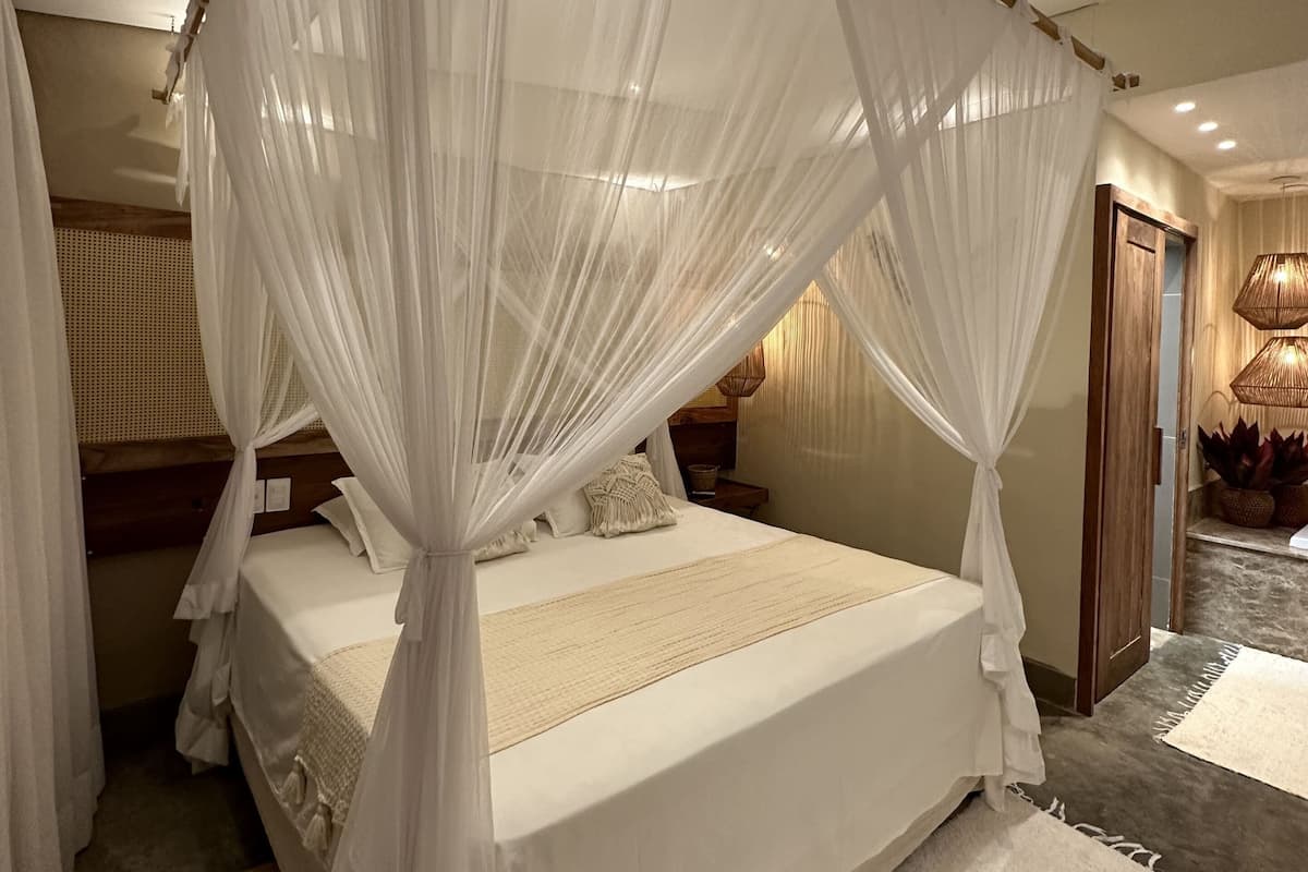 A bedroom with a four-poster canopy bed, a wooden headboard, a bedside table with a lamp, and a view of the hallway.