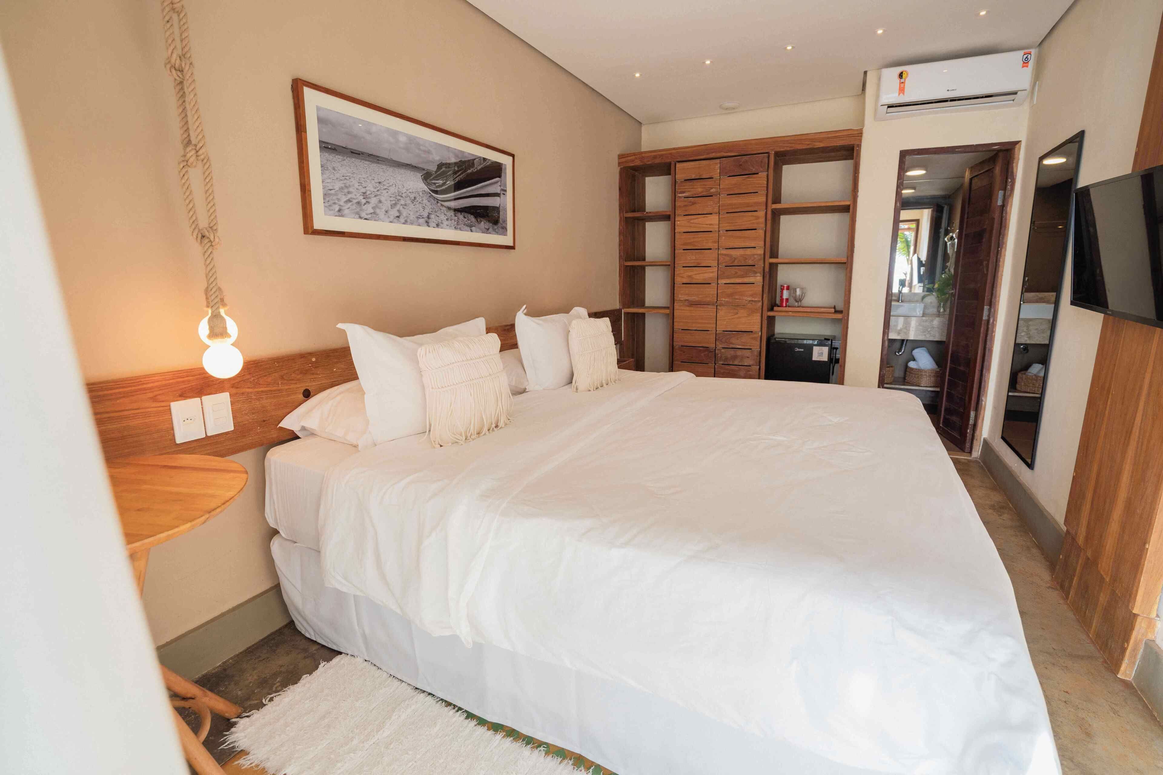 Una cama grande con ropa blanca, un cabecero de madera, una mesita de noche con lámpara, un cuadro enmarcado en la pared y un armario con varios estantes.