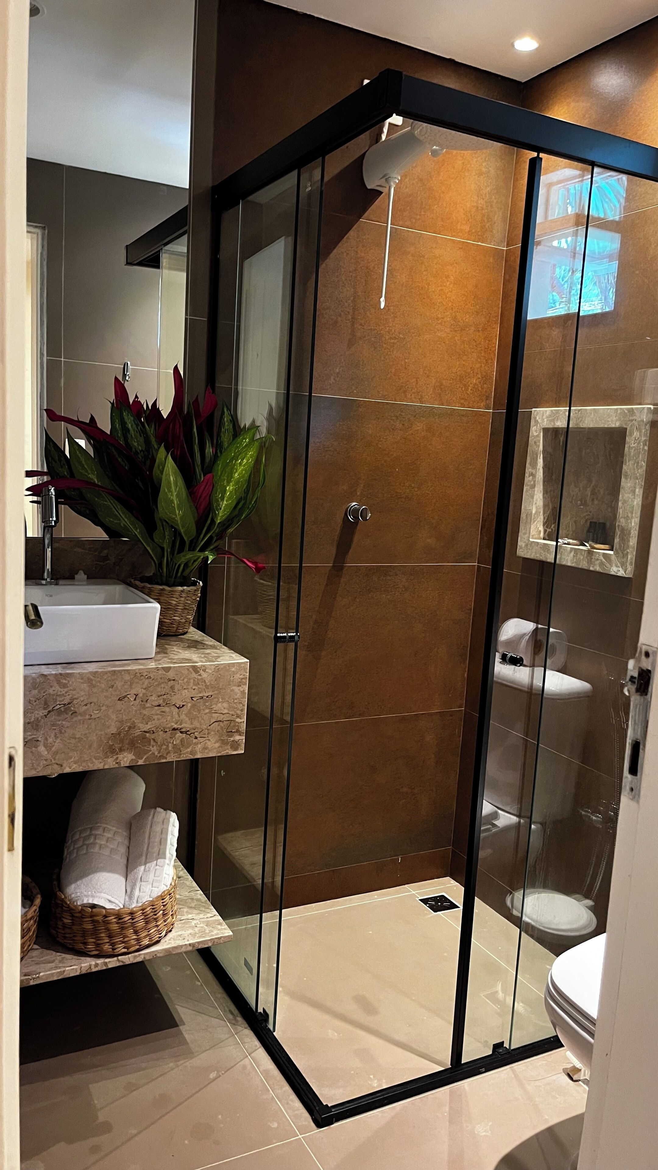 Un baño moderno con ducha envuelta en vidrio, lavamanos con encimera de mármol y una planta en maceta.