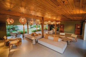Un espacio interior amplio con techo de madera, varios conjuntos de asientos que incluyen sofás y sillones, y lámparas de mimbre colgantes.