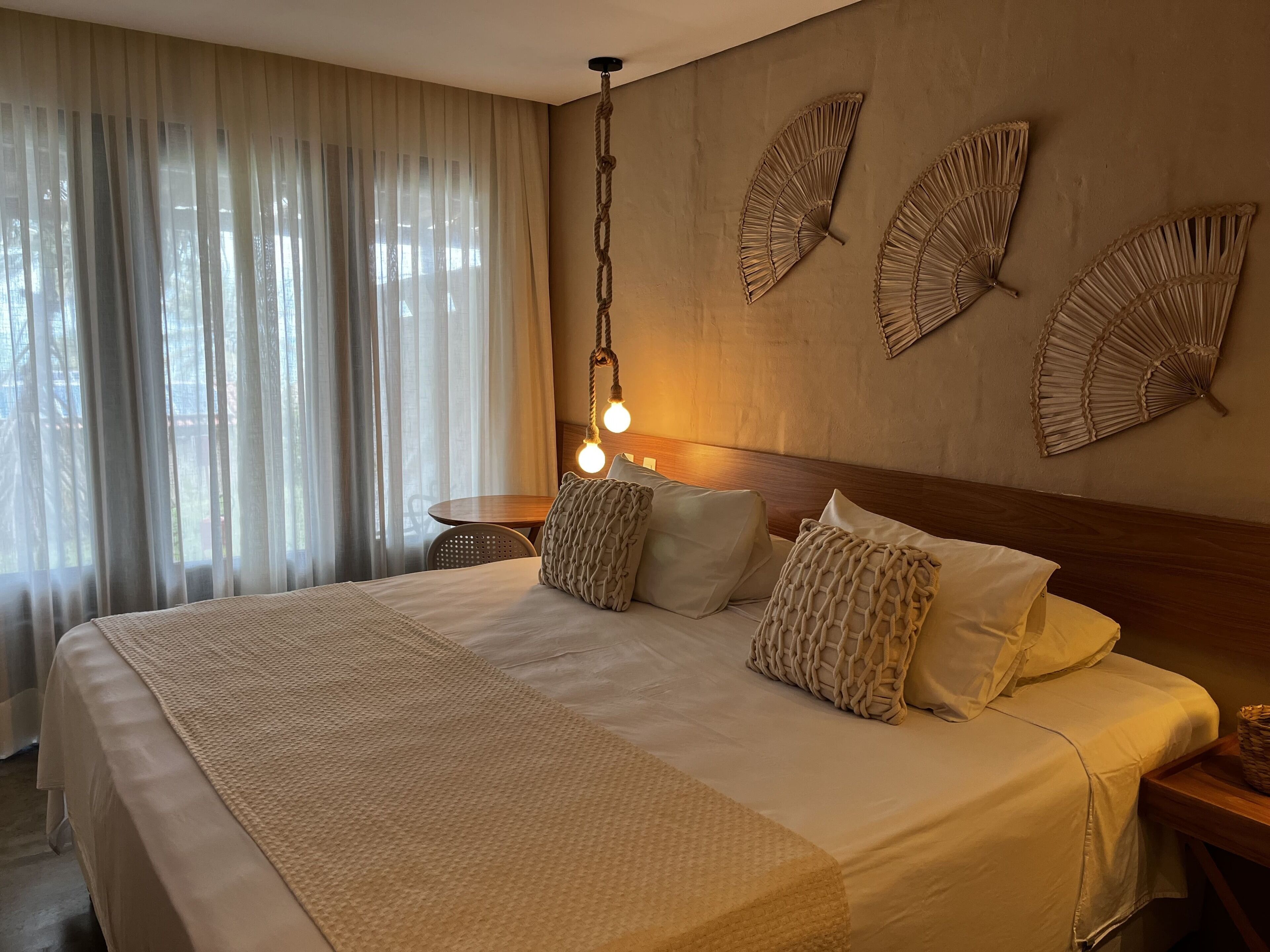 Una cama bien hecha con un edredón texturizado, una mesita de noche de madera y un ventilador fijado a la pared.