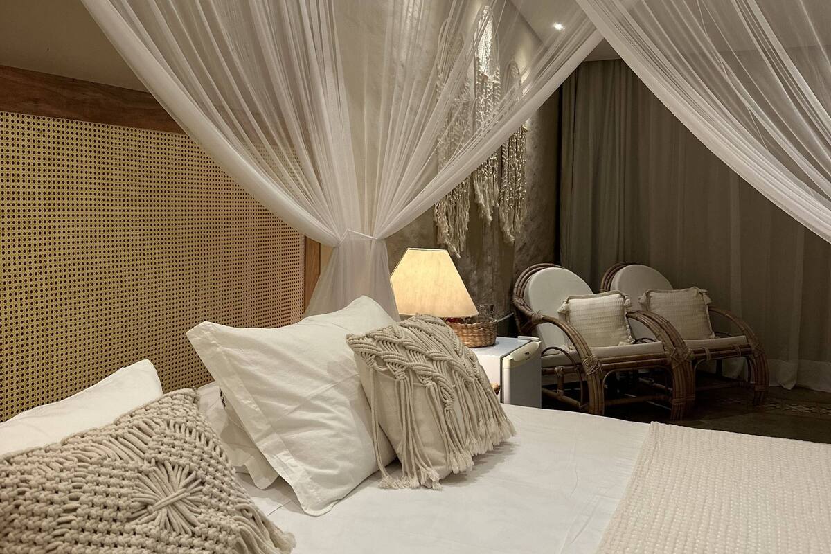 A bedroom with a bed, macrame pillows, a nightstand, a lamp, wicker chairs, and sheer curtains.