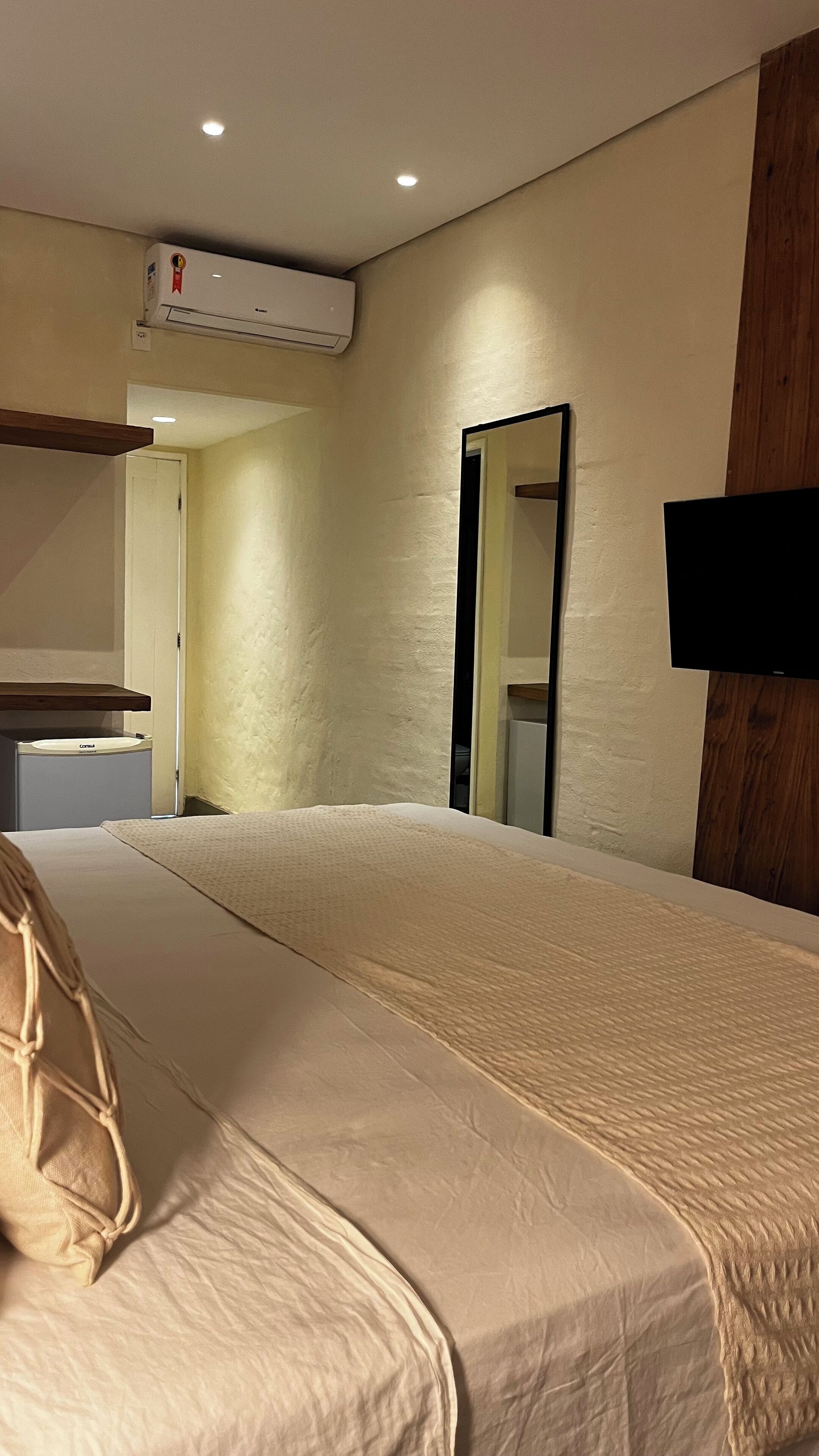 A hotel room with a large bed, a bedside table, a mirror, a television mounted on the wall, and an air conditioning unit.