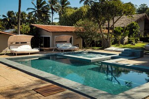 Un área de piscina con sillones de descanso, un jacuzzi y un edificio con techo de paja.