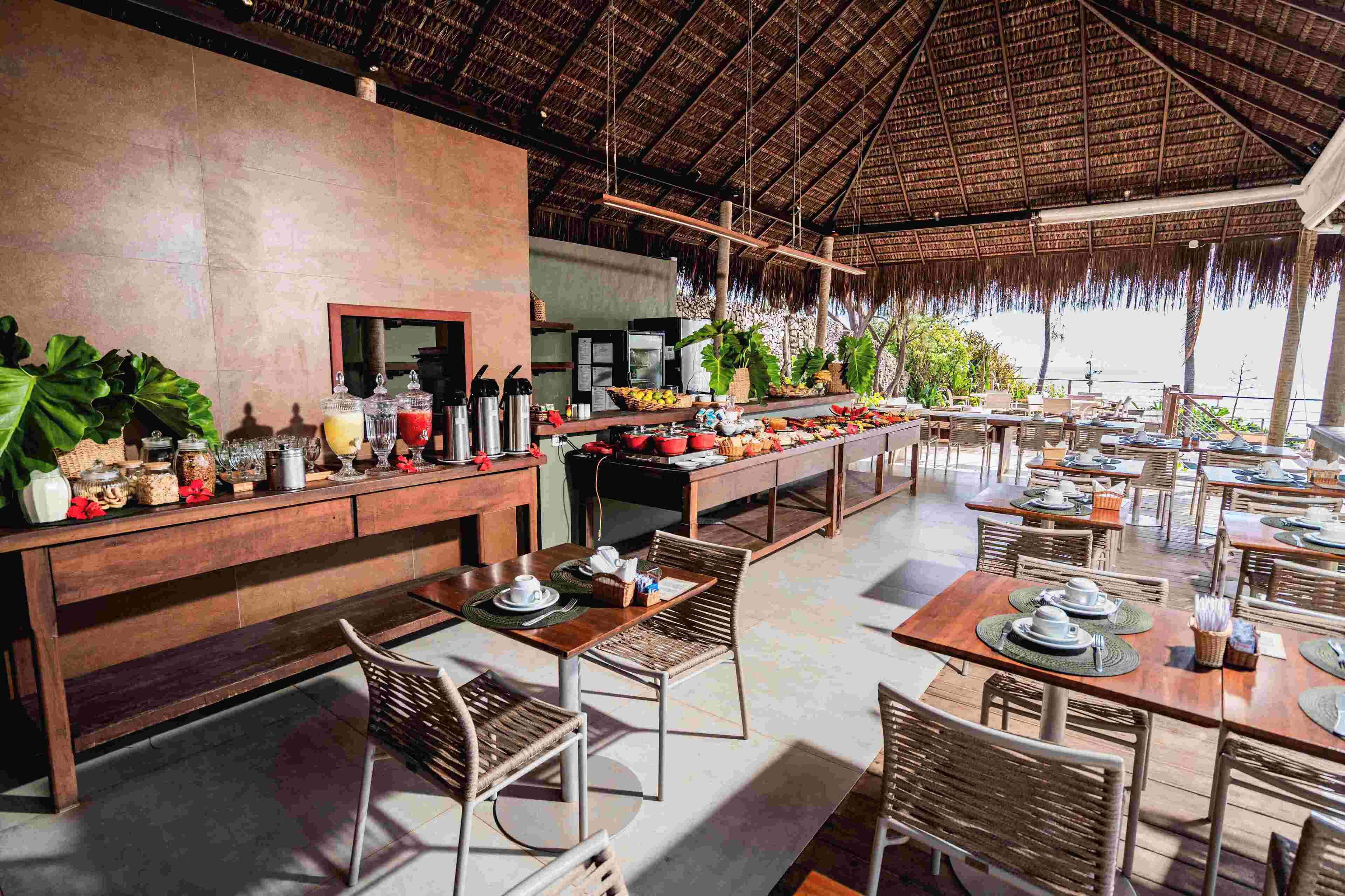 A restaurant with a thatched roof, wooden furniture, and a buffet setup.