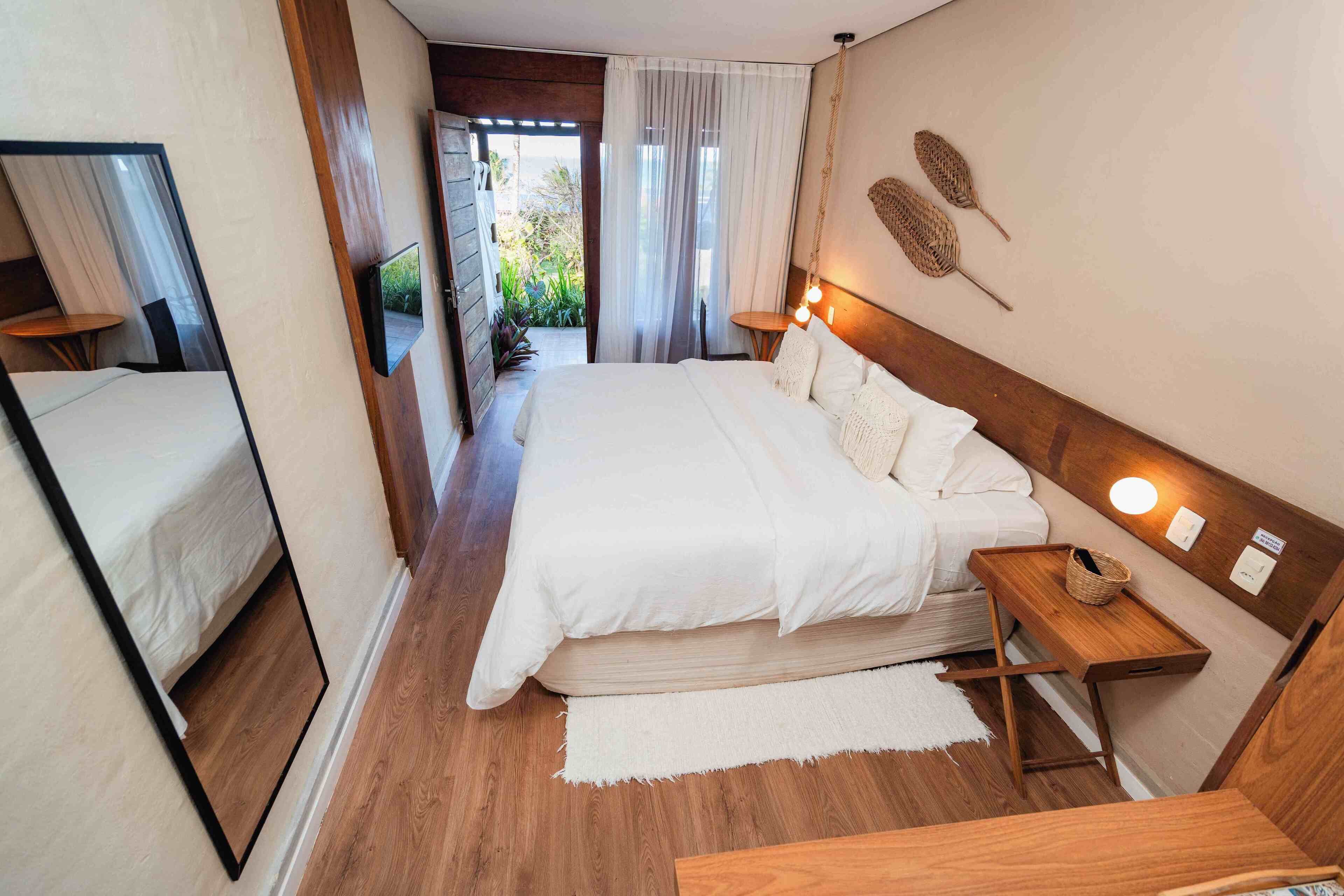 Quarto de hotel com uma cama grande, mesa de cabeceira com abajur, espelho e vista para o exterior.