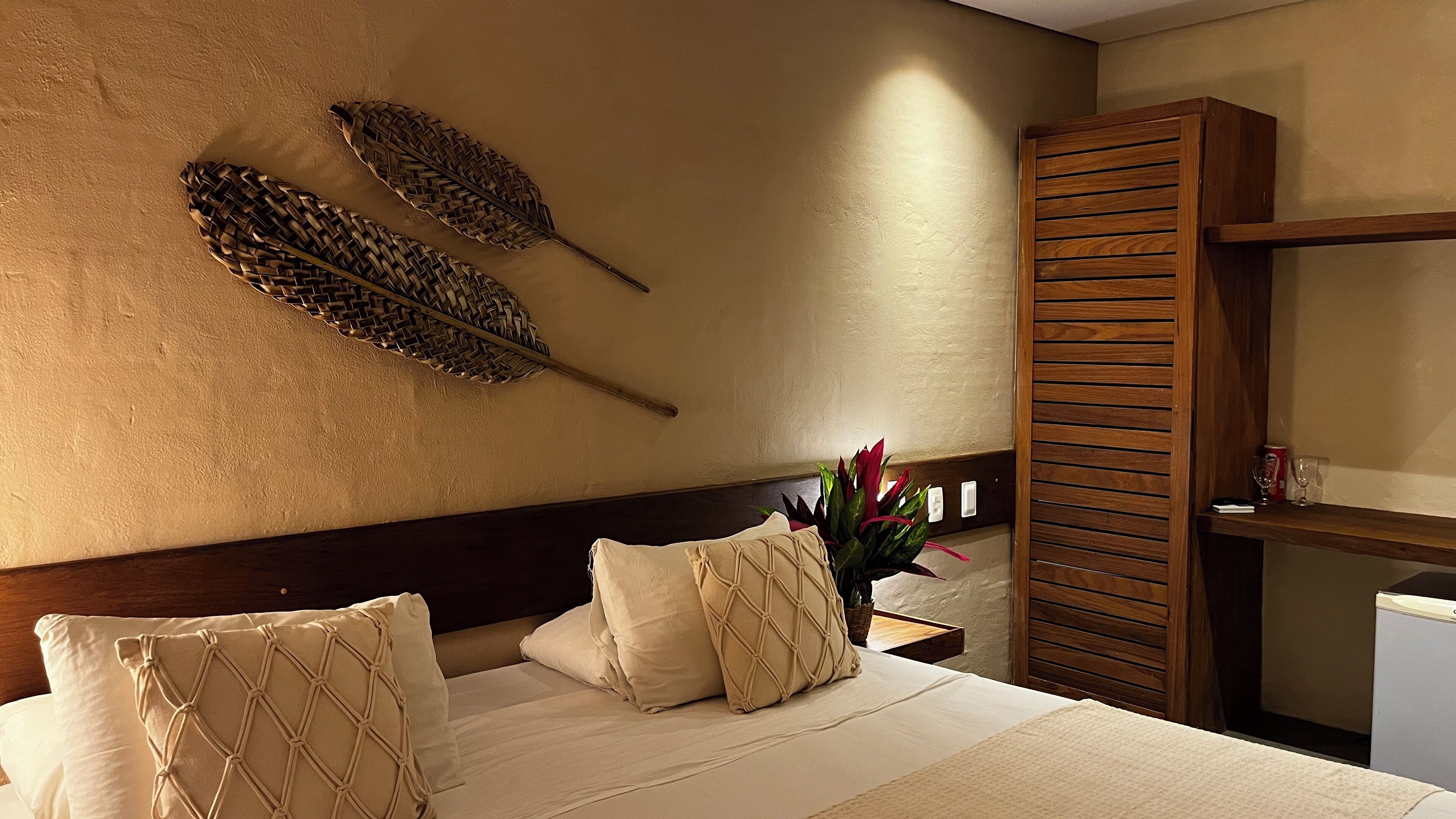 Une chambre d’hôtel avec un lit, deux feuilles décoratives en fibres tressées accrochées au mur, une tête de lit en bois et une table de chevet sur laquelle est posé un vase de fleurs.