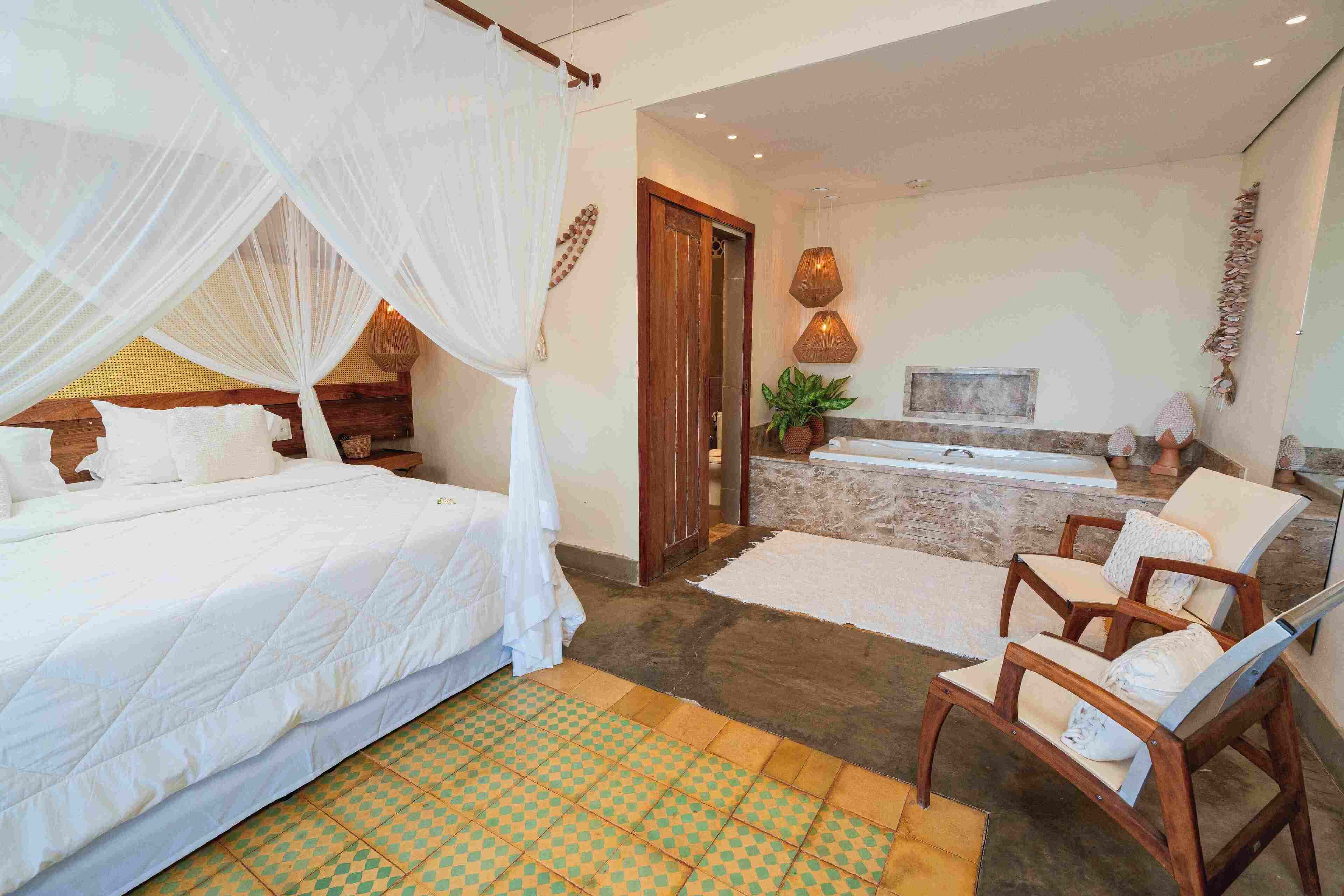 Quarto com cama, área de estar com cadeiras, uma janela grande e banheiro com banheira.