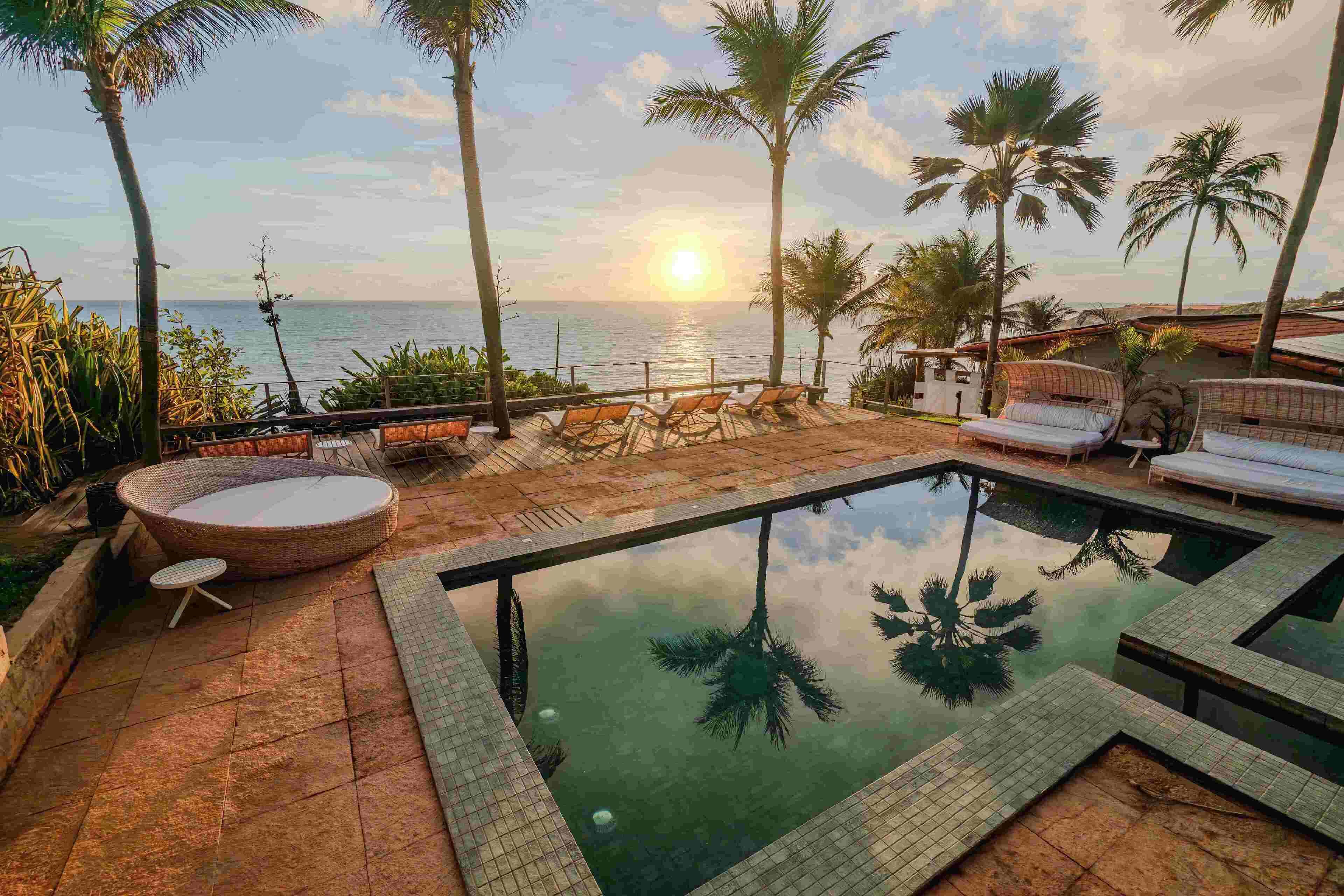 A pool area with lounge chairs and a hot tub, surrounded by palm trees and a view of the ocean at sunset.
