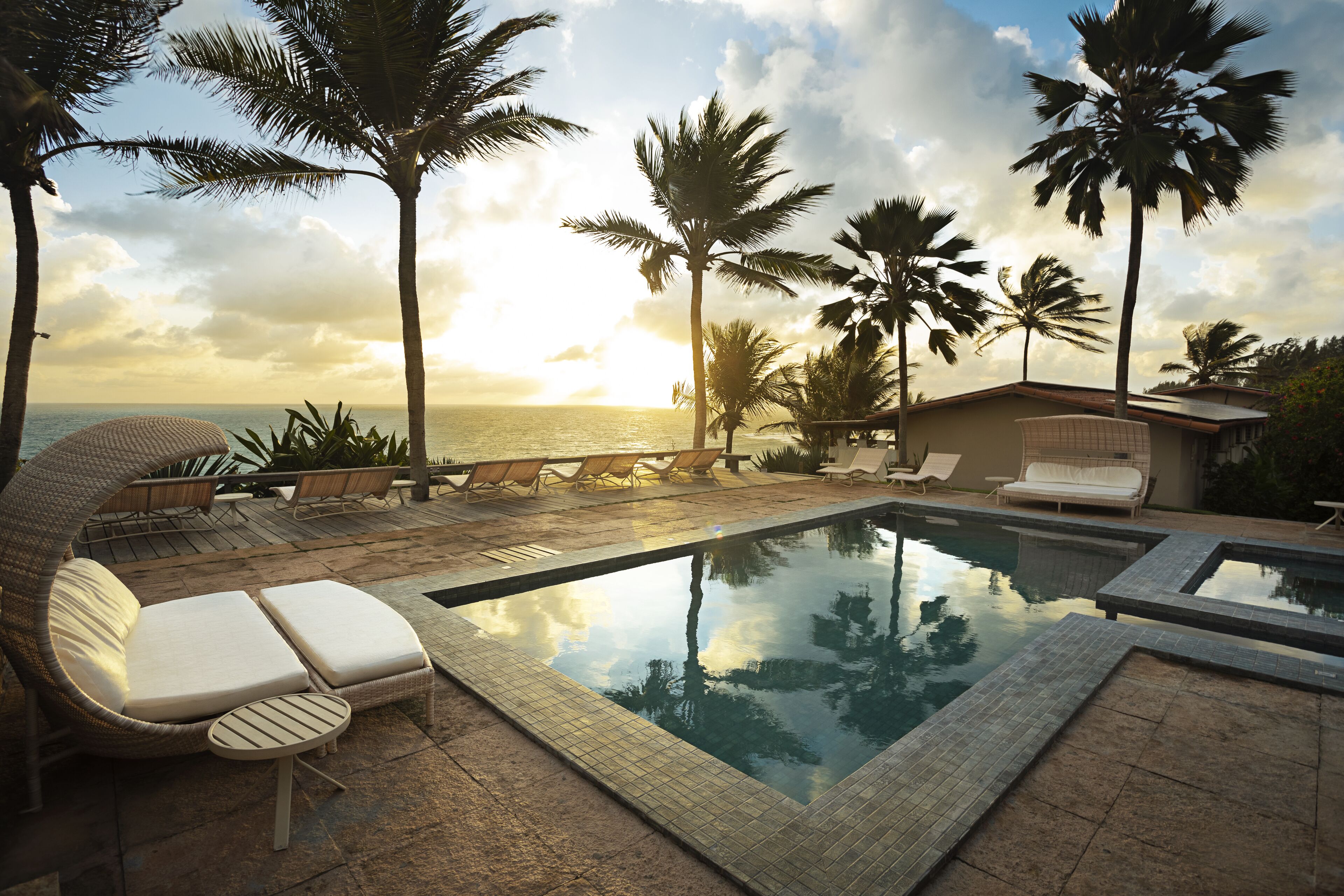 A poolside area with lounge chairs, a small table, and a view of the ocean at sunset.