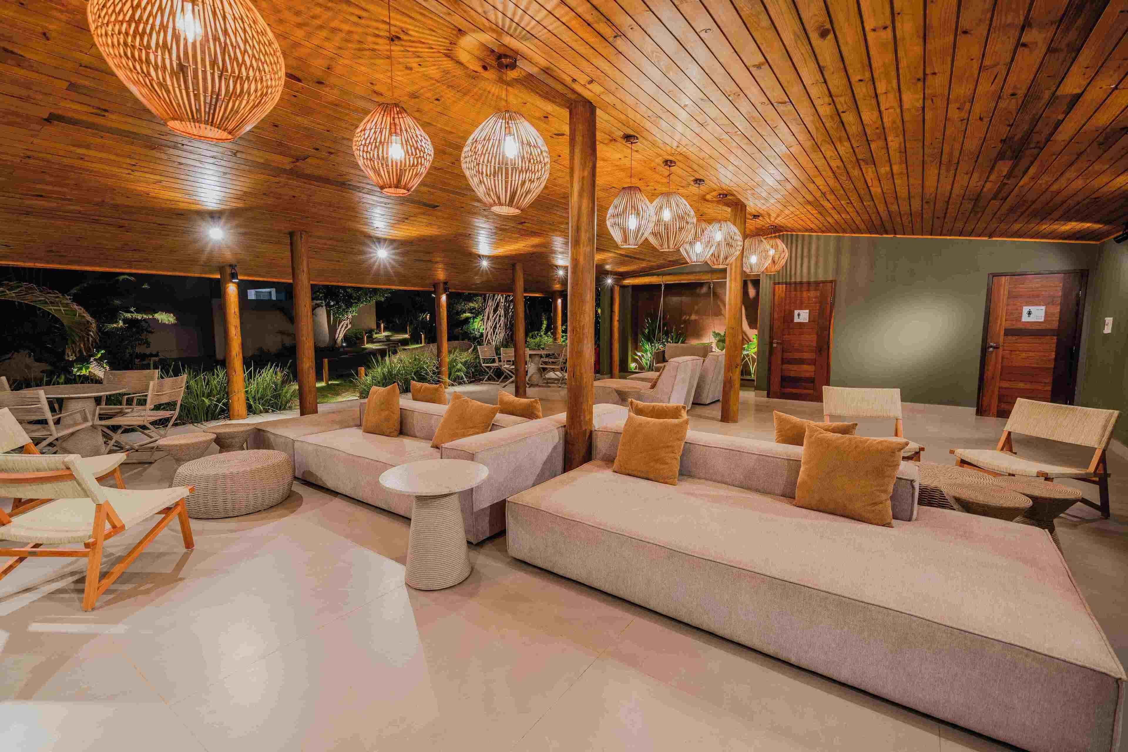 A spacious indoor lounge with wooden ceiling and hanging light fixtures, featuring a large sectional sofa, cushioned chairs, and a round table.