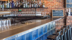 A bar with a brick wall, hanging pendant lights, a clock, and a menu board. - Hotel Belley (Québec City)