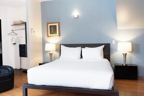 A hotel room with a bed, two bedside lamps, a framed picture on the wall, a coat rack, and a black chair. - Hotel Belley (Québec City)