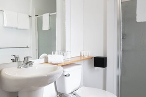 Un baño con lavabo blanco, espejo y váter. Hay un perchero sobre el lavabo y un estante con artículos de aseo.