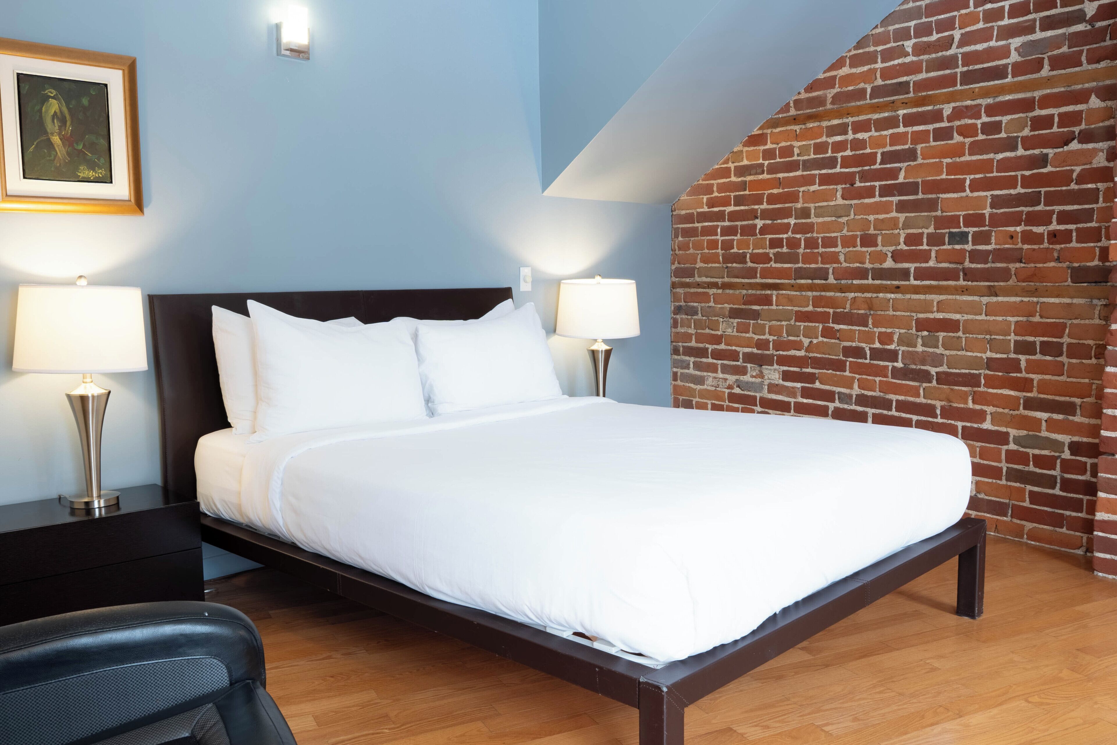 A neatly made bed with white linens, two bedside lamps, a framed picture on the wall, and a brick wall adjacent to the bed.