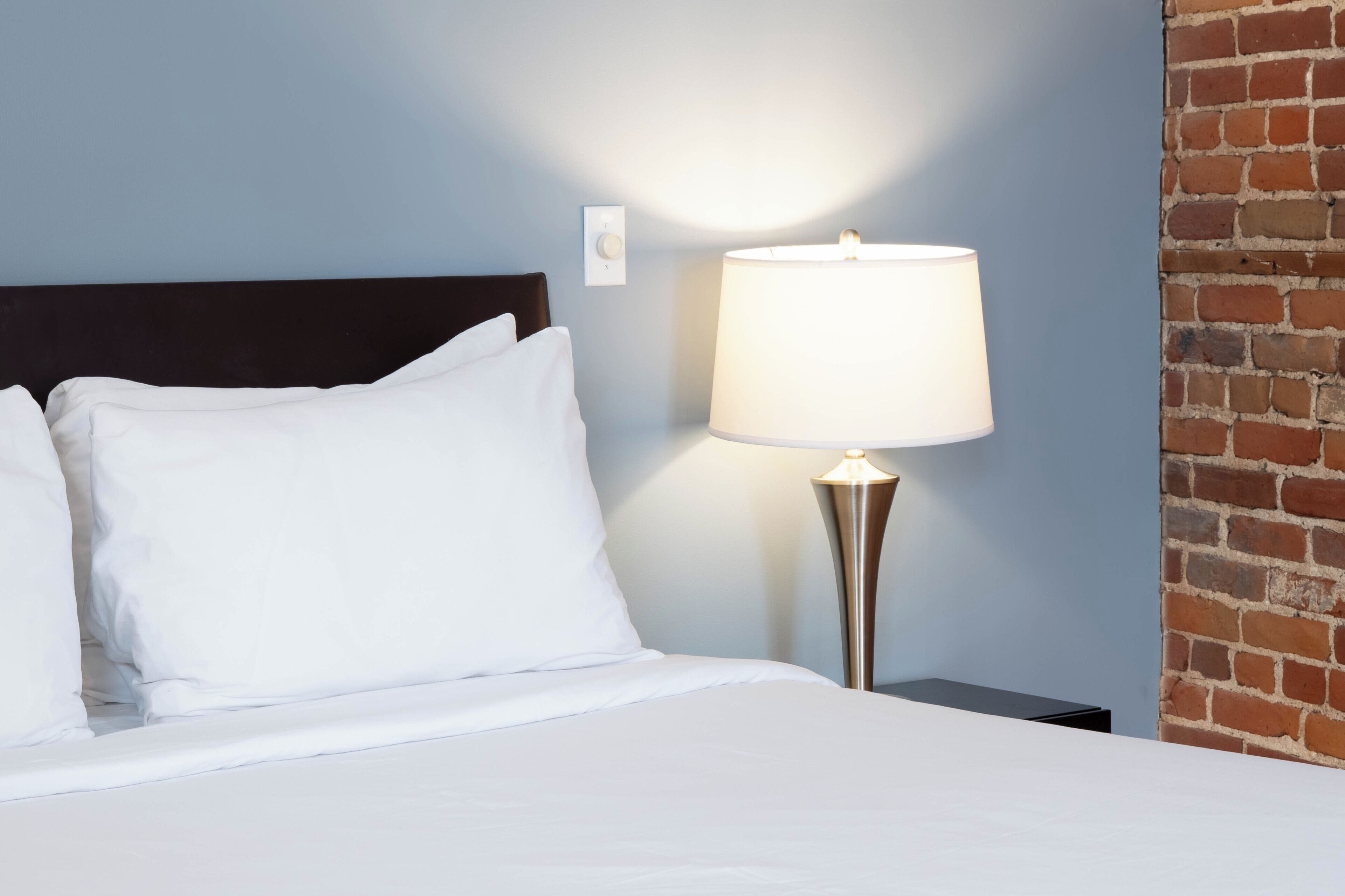 A neatly made bed with white linens, a modern lamp with a white shade, and a brick wall.