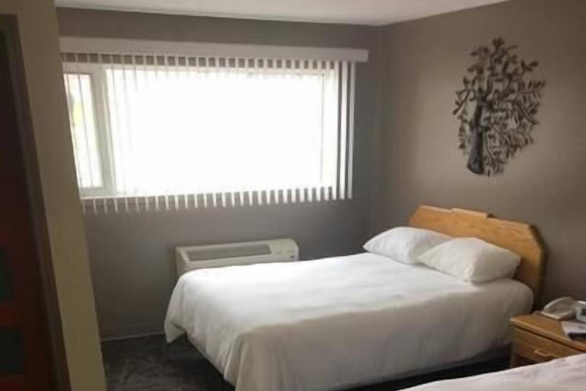 Hotellihuone, jossa on kaksi sänkyä, patteri, puhelin yöpöydällä ja ikkuna, jossa on sälekaihtimet.