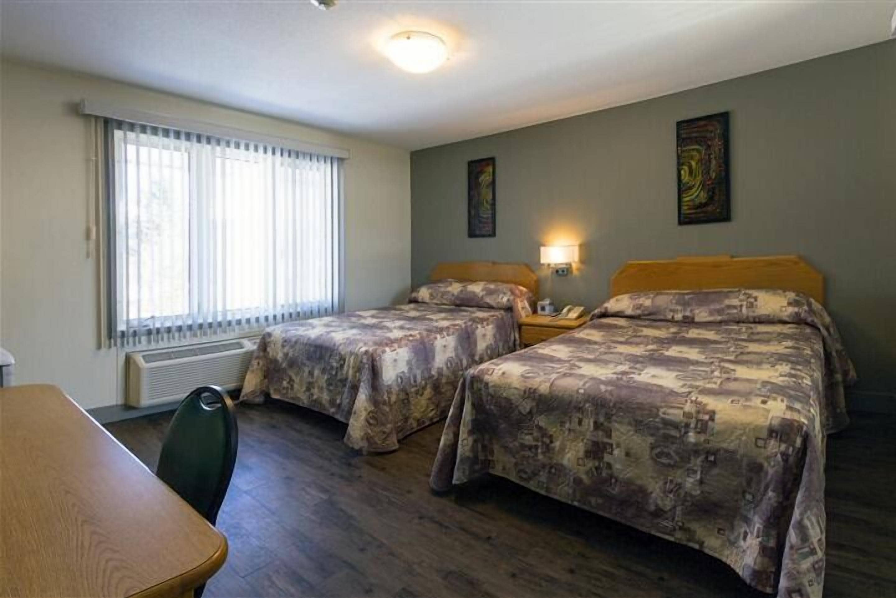 Una habitación con dos camas, una mesa y una silla. Cuenta con una ventana con persianas, un calentador y dos lámparas fijadas a la pared.