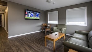 Una sala de estar con televisor de pantalla plana, una mesa de centro de madera, un sofá verde y dos ventanas con persianas.