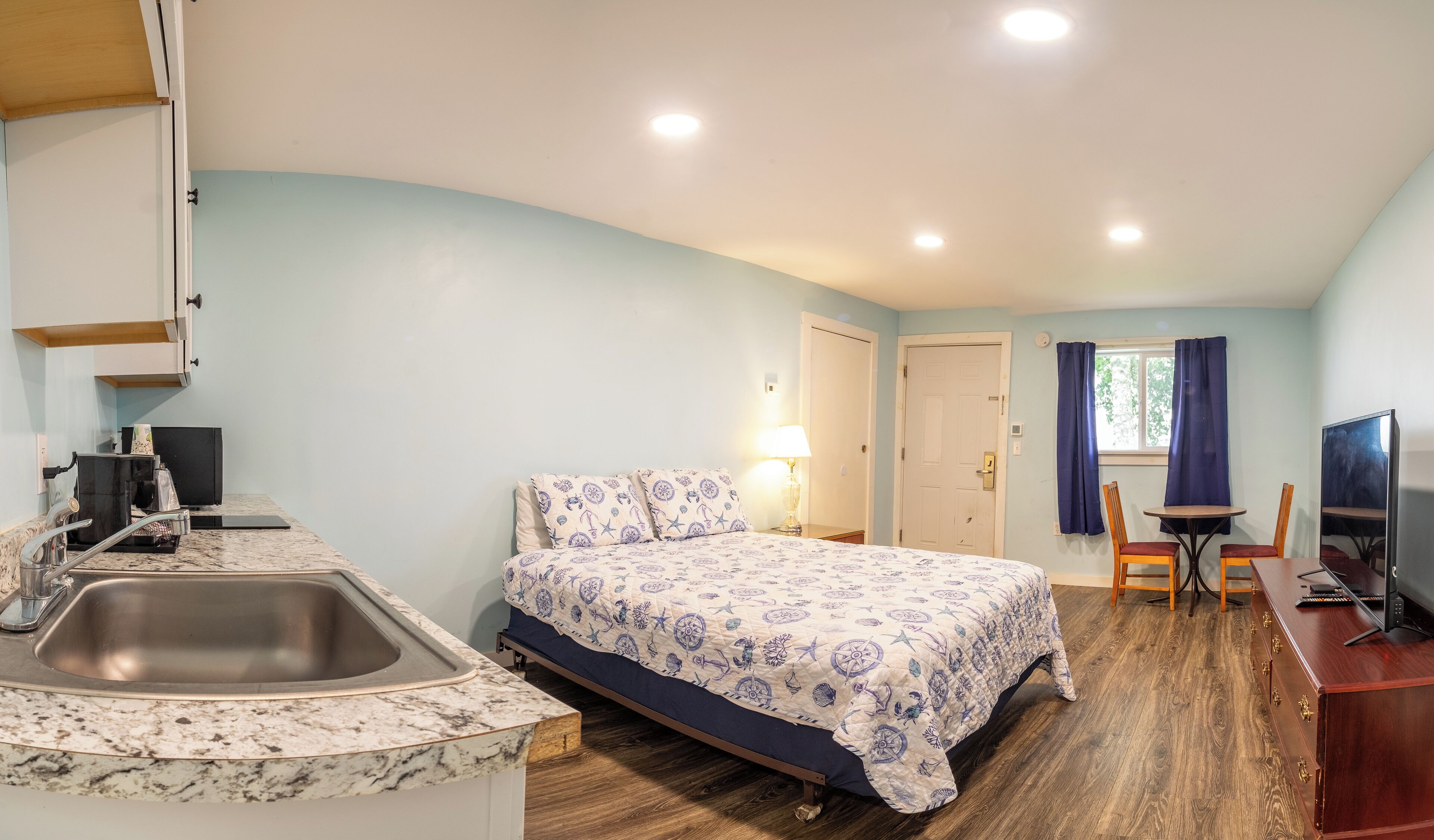 A compact hotel room with a bed, a desk, a chair, a TV, a sink, and a window with curtains.