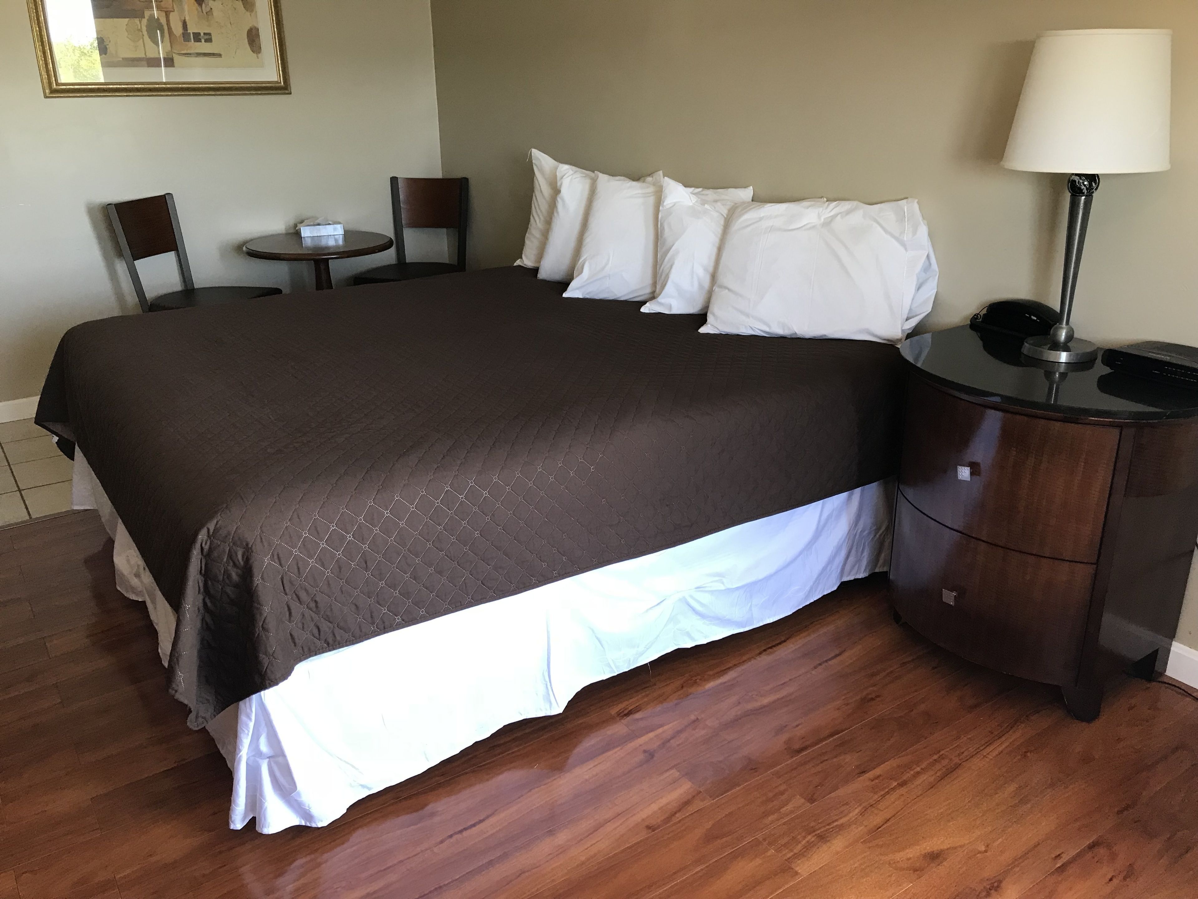 A neatly made bed with a dark quilt, a bedside table with a lamp, and a small round table with a chair.