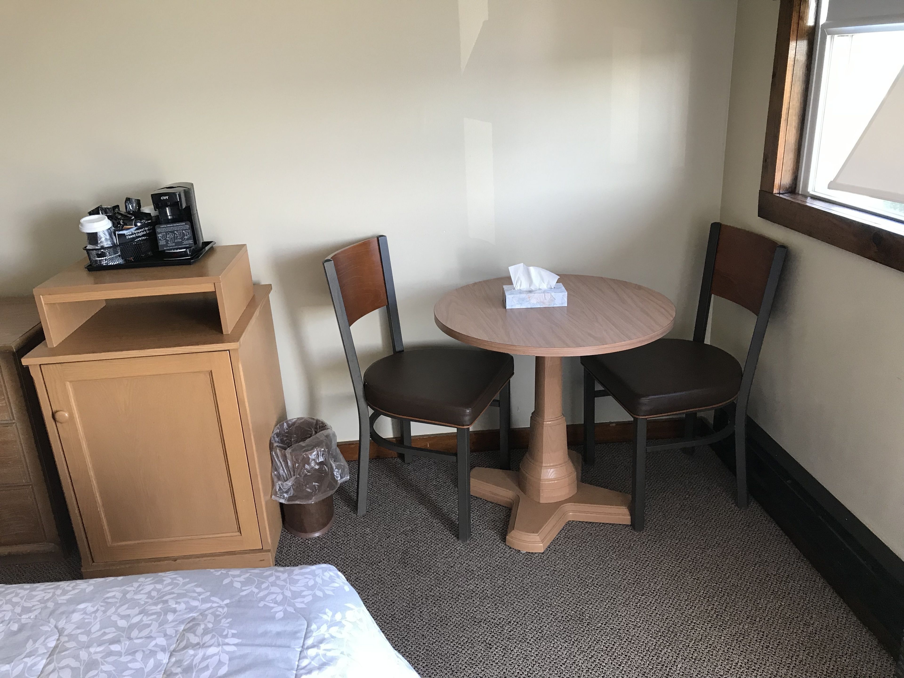 A small hotel room with a bed, a wooden cabinet, a round table with two chairs, a trash can, and a window with blinds.