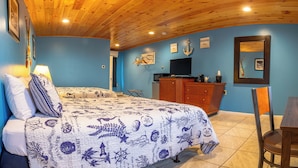 Una habitación con techo de madera, una cama con colcha azul y blanca estampada, un escritorio con silla y un televisor sobre un mueble de madera.
