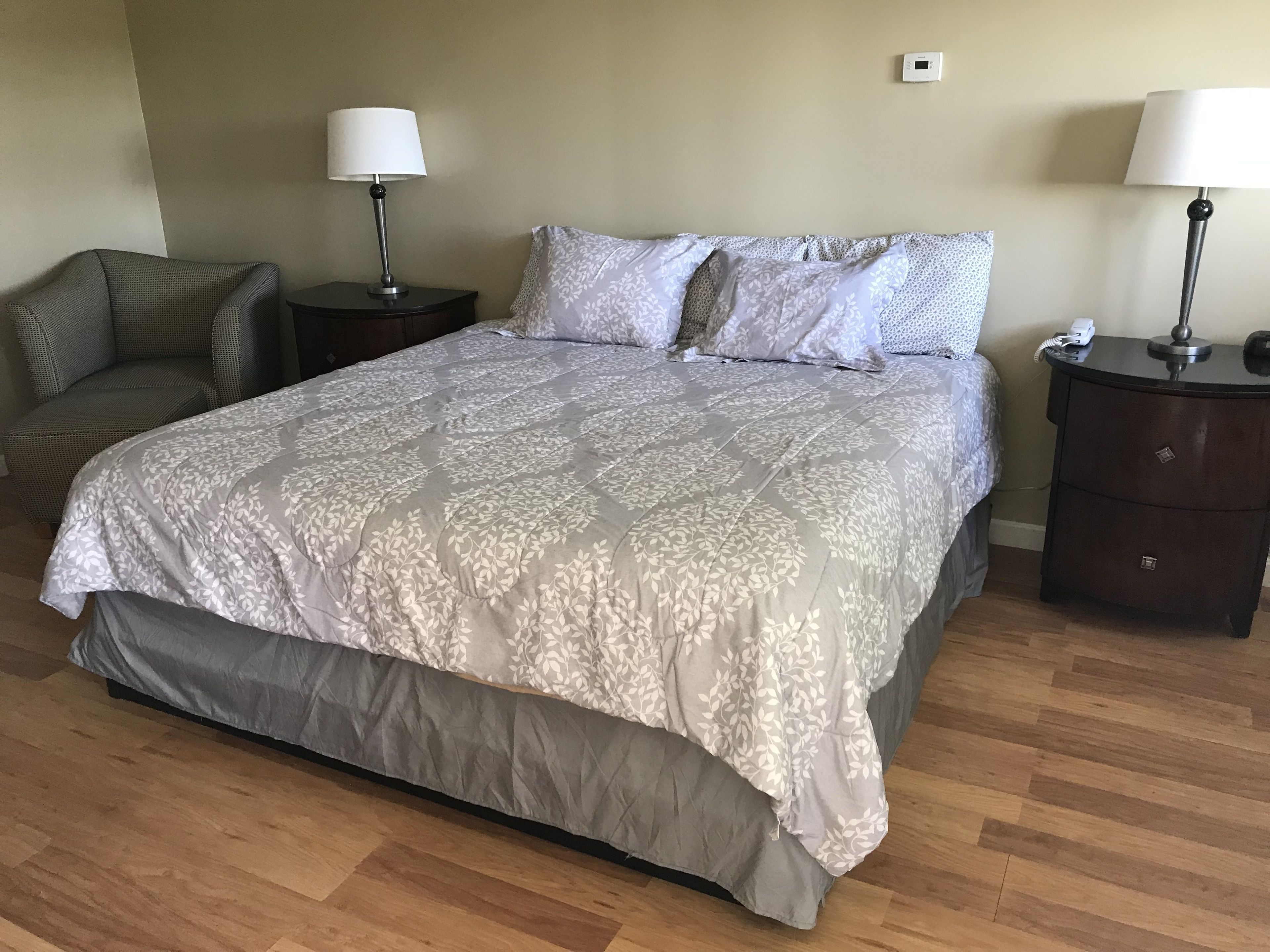 A bedroom with a large bed, two nightstands, a chair, and a wall-mounted thermostat.