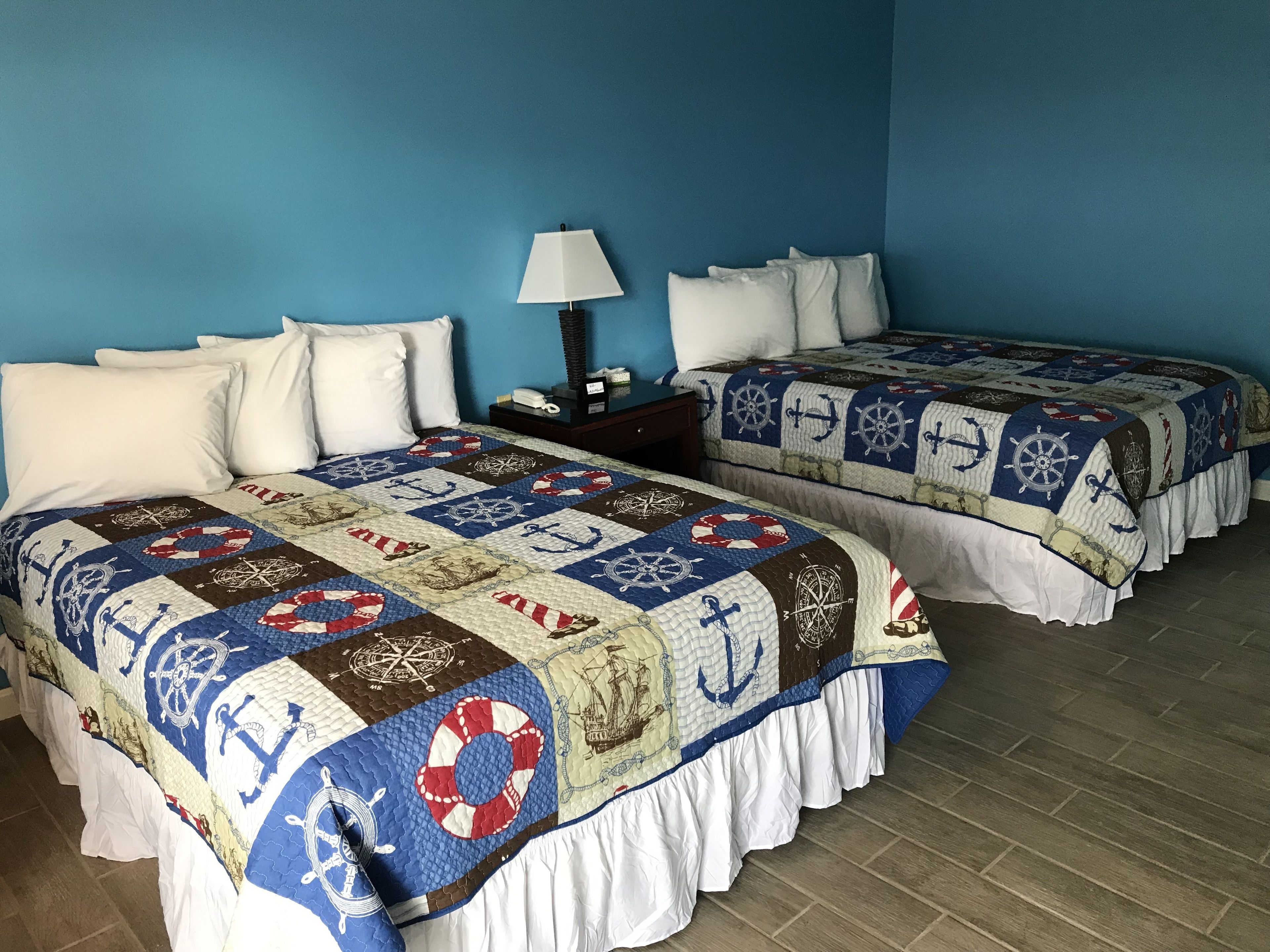 Two beds with nautical-themed quilts, a nightstand with a lamp, and a blue wall.